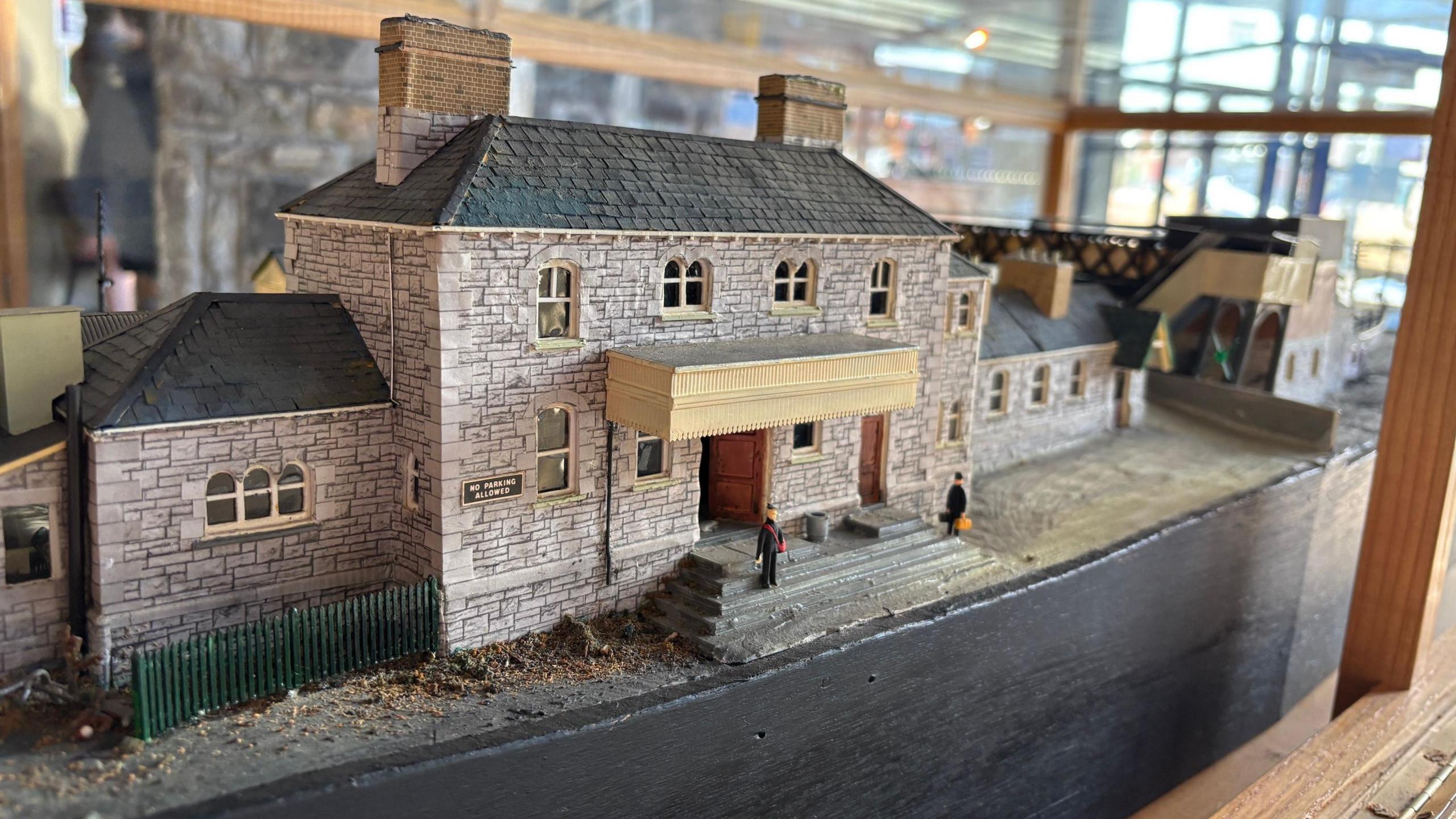 A model railway station sits inside a wood and glass display case. The model biggest in shot is of the station building a grey brick structure with a tiled roof.