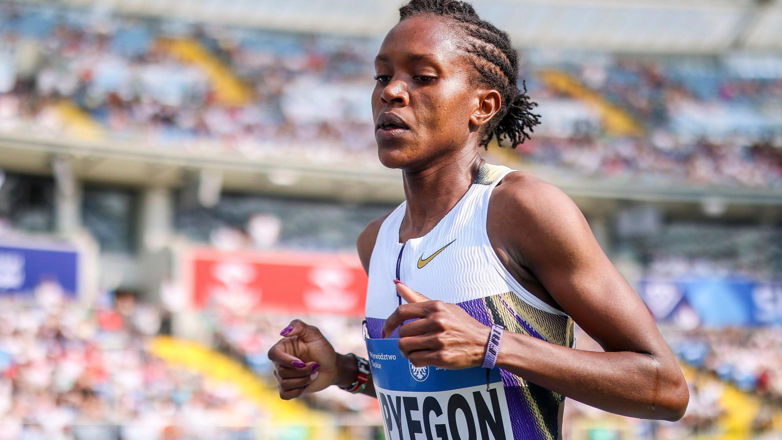Faith Kipyegon seen from stomach up casts her eyes down while running a track race in a white and purple vest, with a packed crowd seen out of focus in the distance behind her