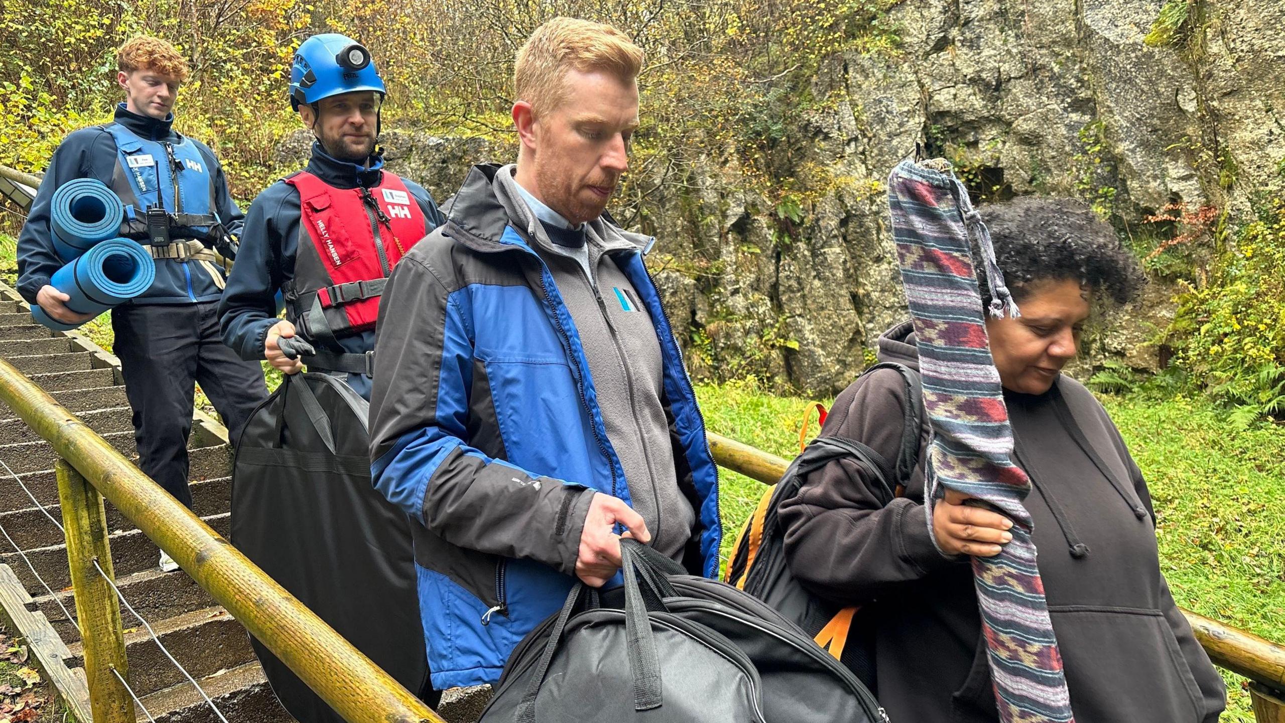 A group of people walk down an outdoor staircase. They are in a woodland, and they are carrying instruments. one person's wearing a hard hat with a torch another is carrying yoga mats.