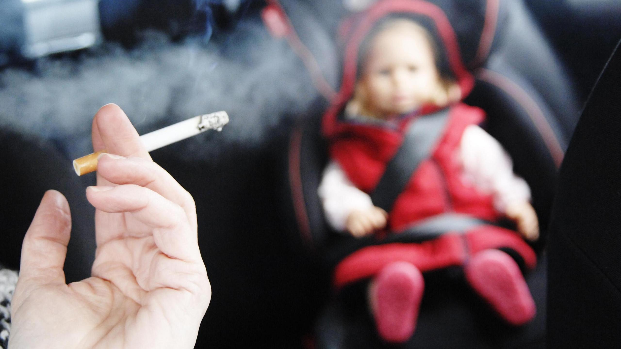 A toddler in a red coat sits in a safety seat in the back of a car, while someone in the front seat smokes.