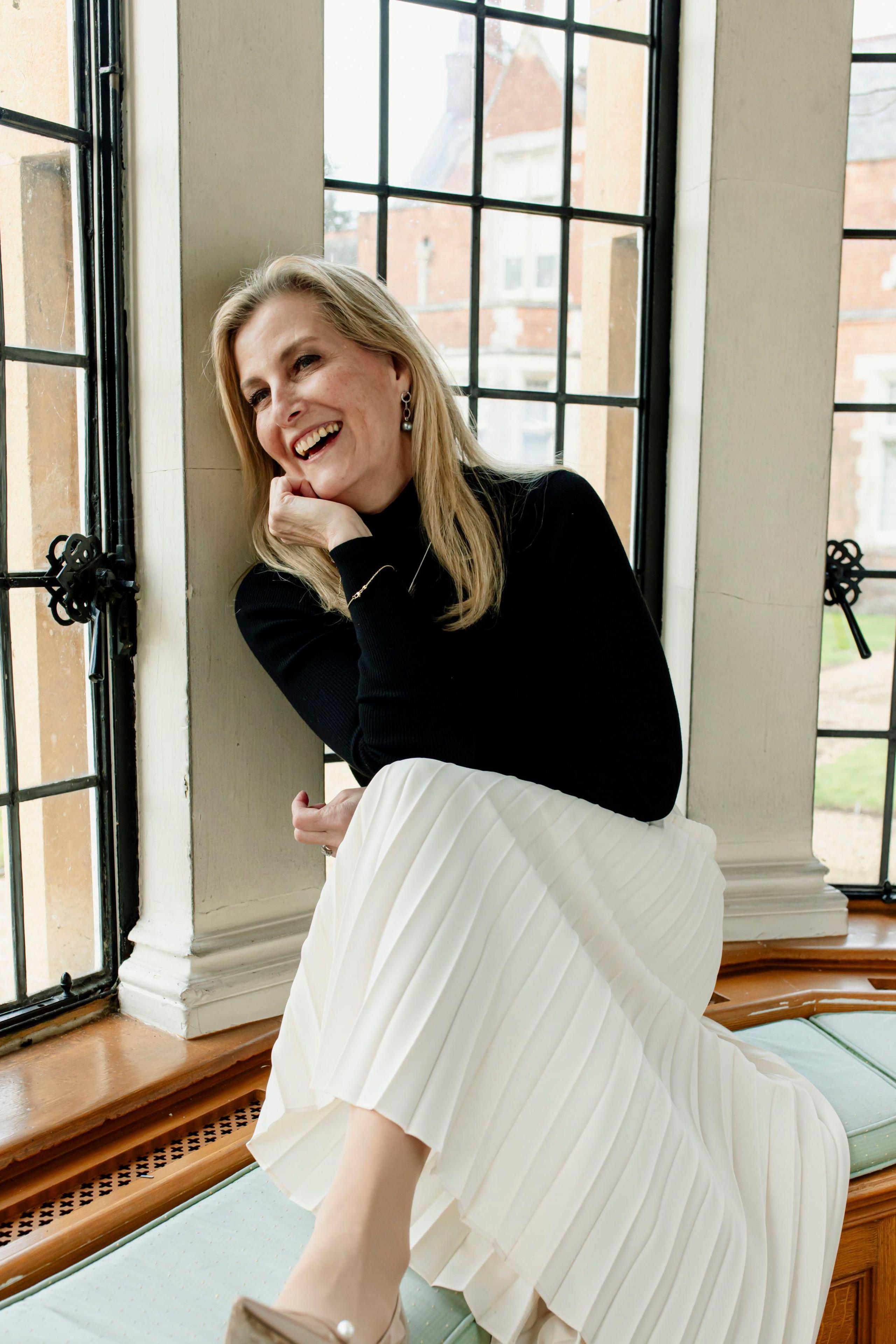 Sophie, who has long blonde hair and wears a black turtleneck with a pleated cream midi skirt, looks relaxed as she sits on a cushioned bench in a bay window and laughs while cupping her face with her hand 