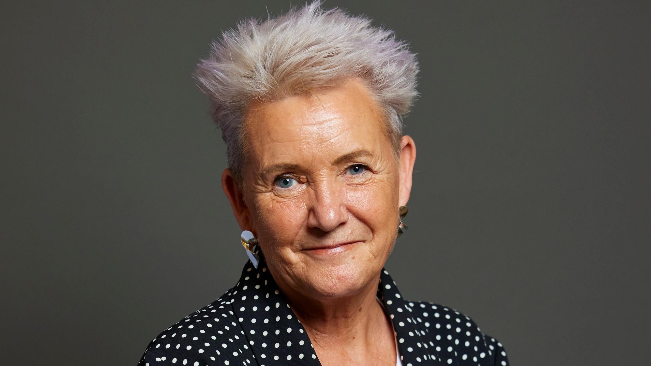 Official portrait of Lorraine Beavers, Blackpool North and Fleetwood MP, taken against a grey backdrop. Lorraine has short white hair, is wearing silver earrings, and a black and white polka dot outfit. She is smiling.