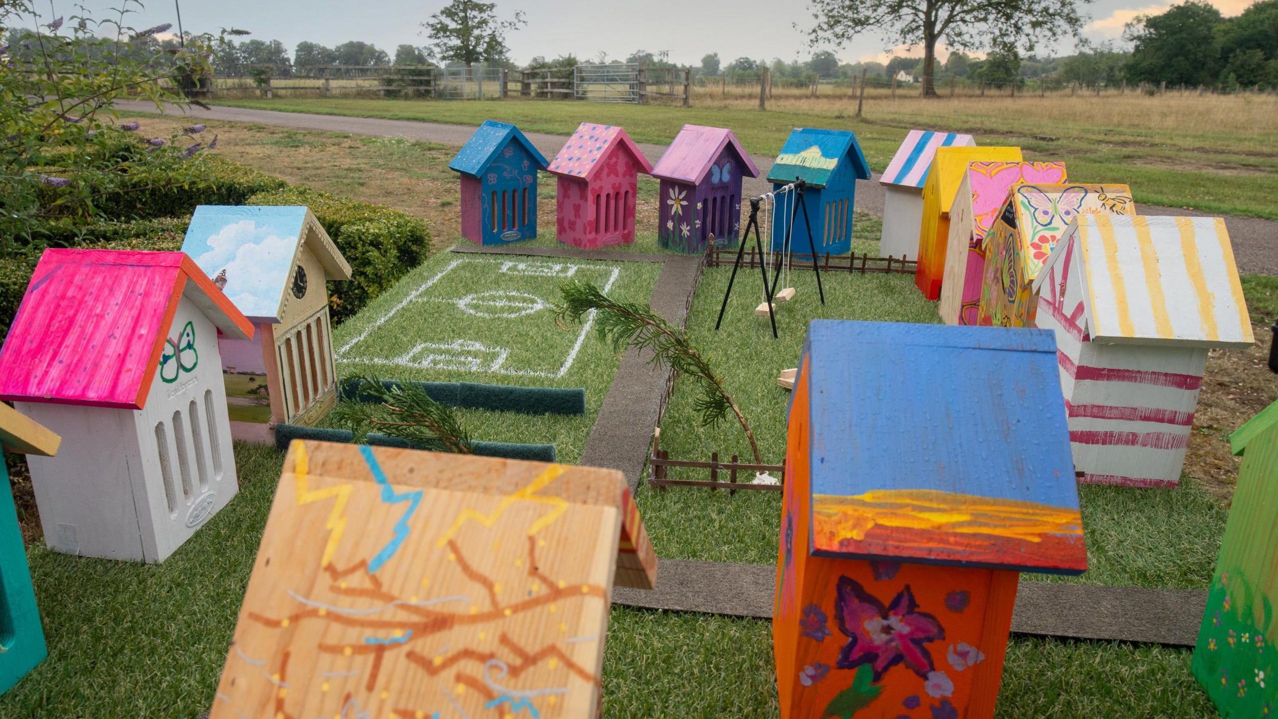 Fourteen brightly coloured small wooden houses are arranged on artificial grass around a pretend football pitch, with fields and trees in the background.
