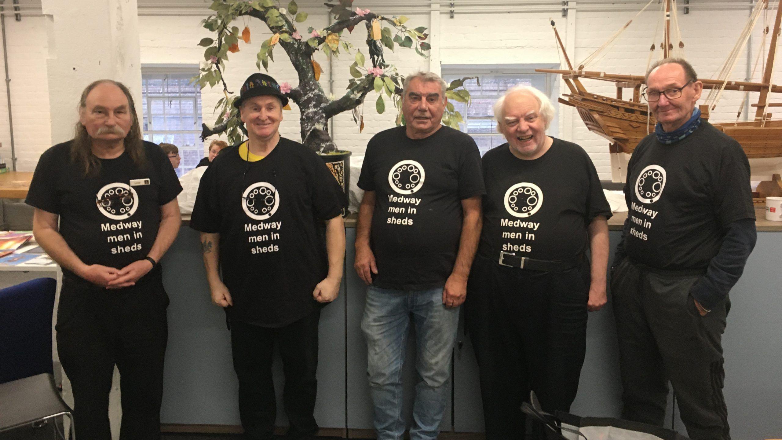 Five men pose for a photograph wearing black Medway Men In Sheds t-shirts in their workshop. A wooden model of a sailing ship is behind them.