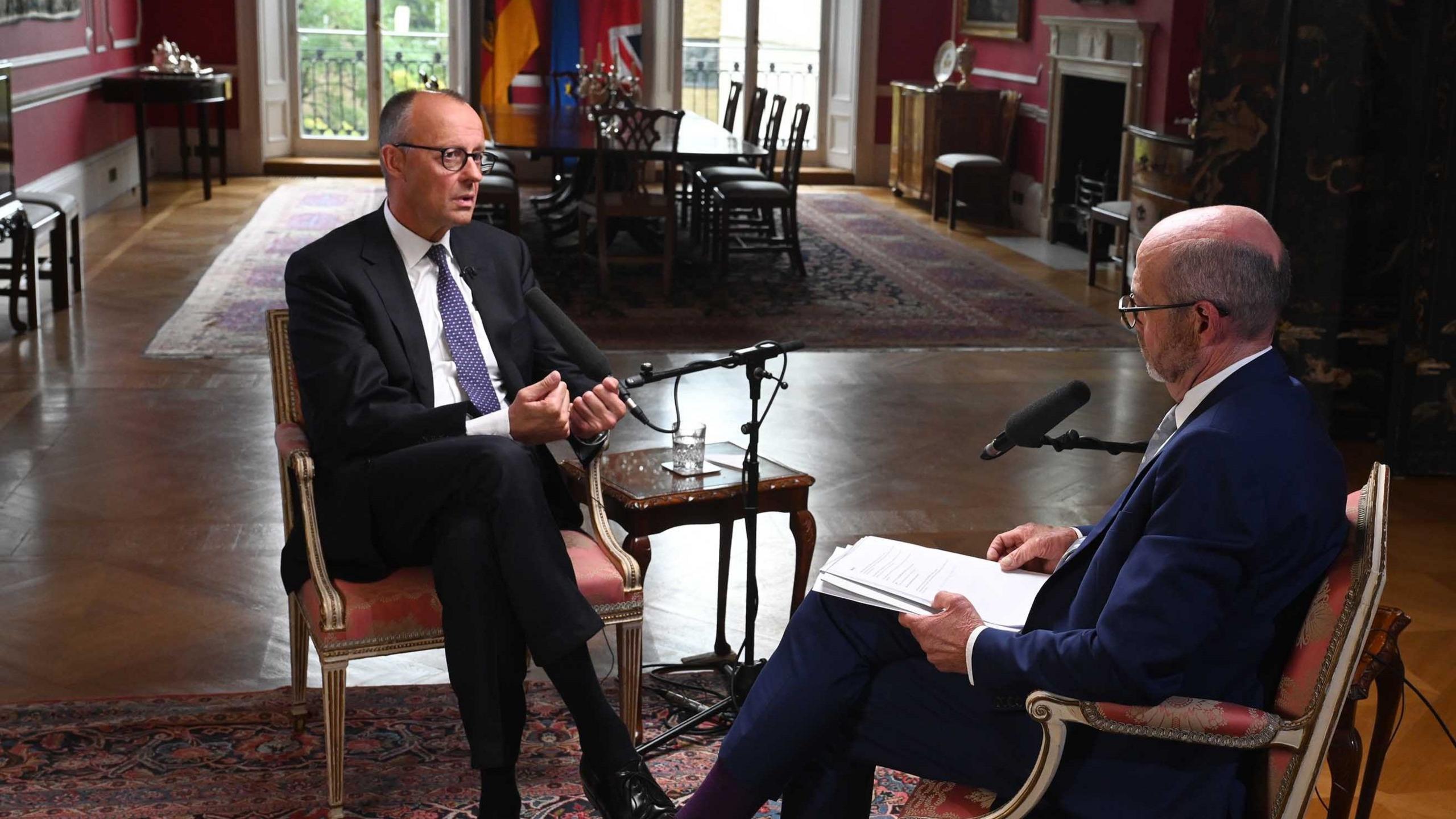 Germany's chancellor sits with his legs crossed on the left in a suit, talking to the BBC's Nick Robinson on the right who has papers on his lap