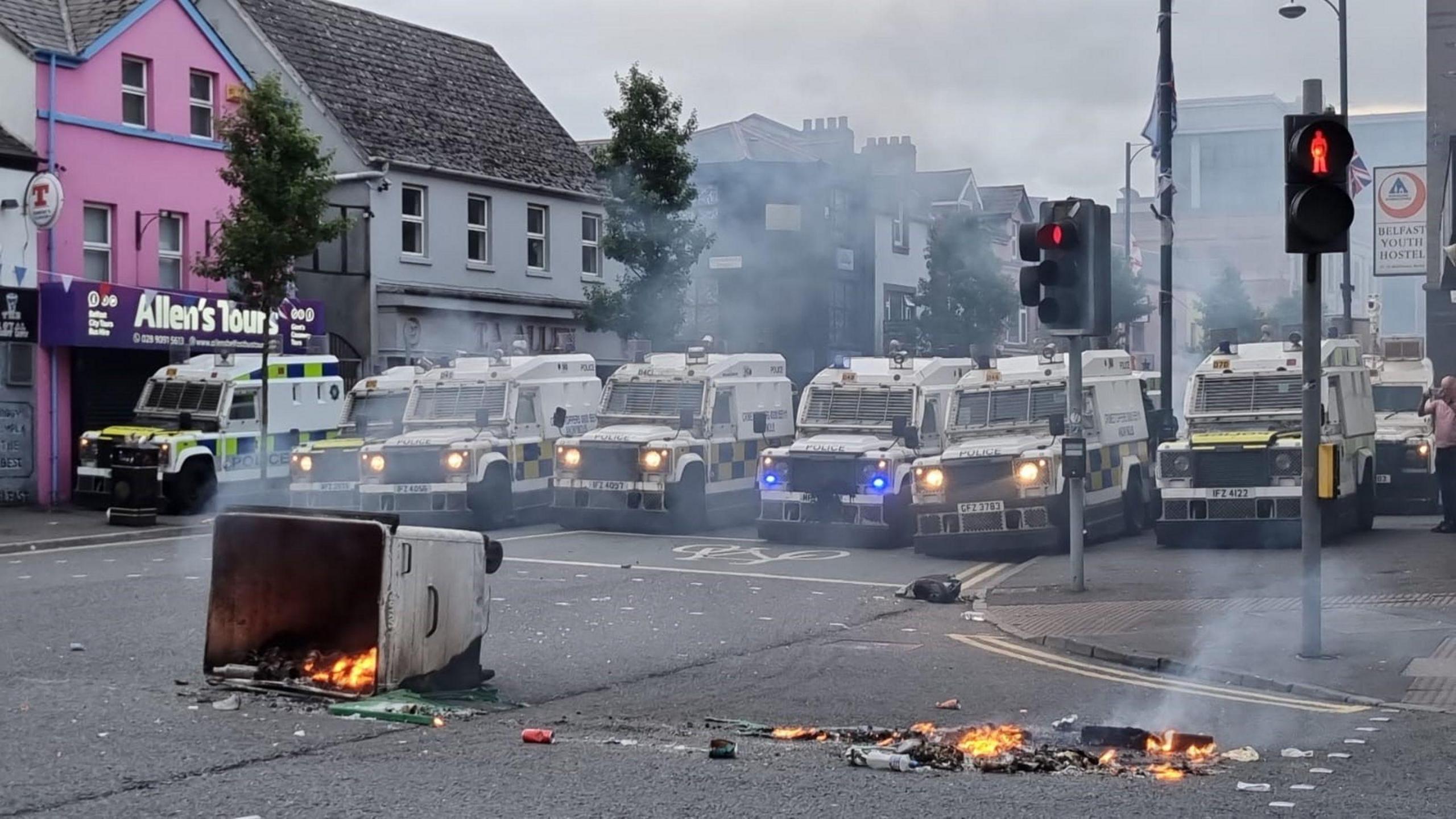 PSNI vans lined up in a row on a road in Belfast with a bin on fire in front of them