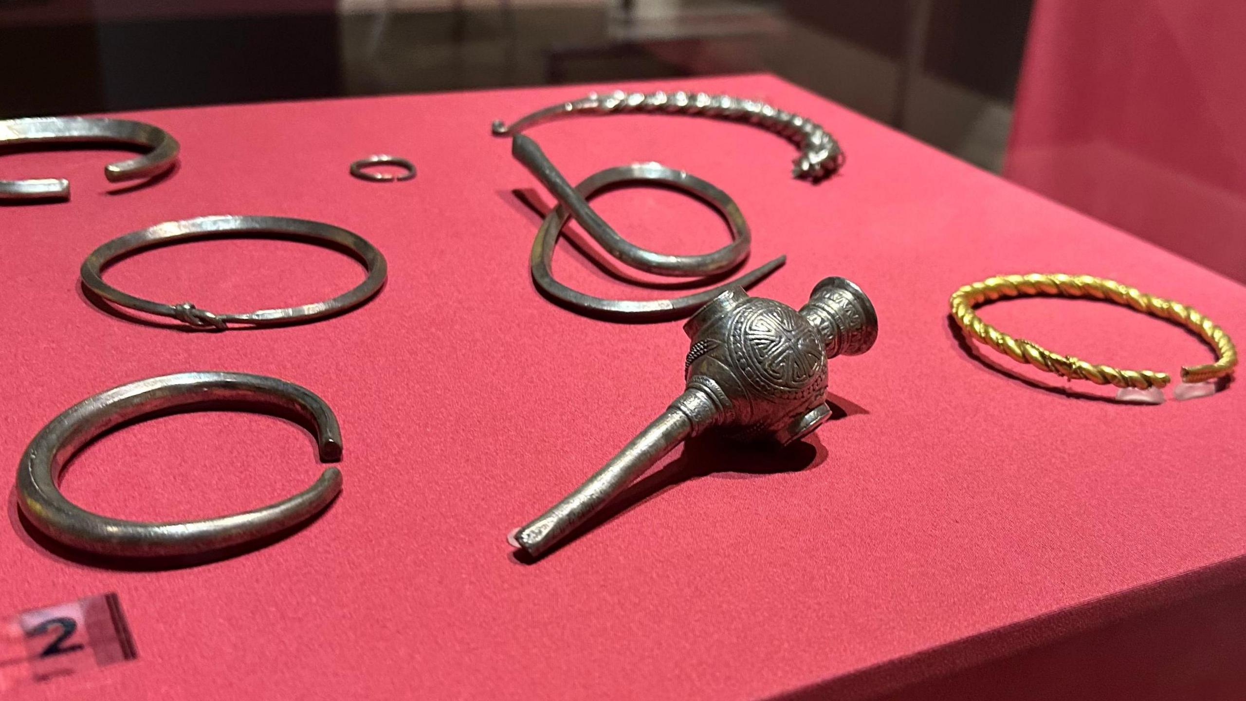 Silver arm bands and other items and a gold bracelet placed on a red box in a glass case.