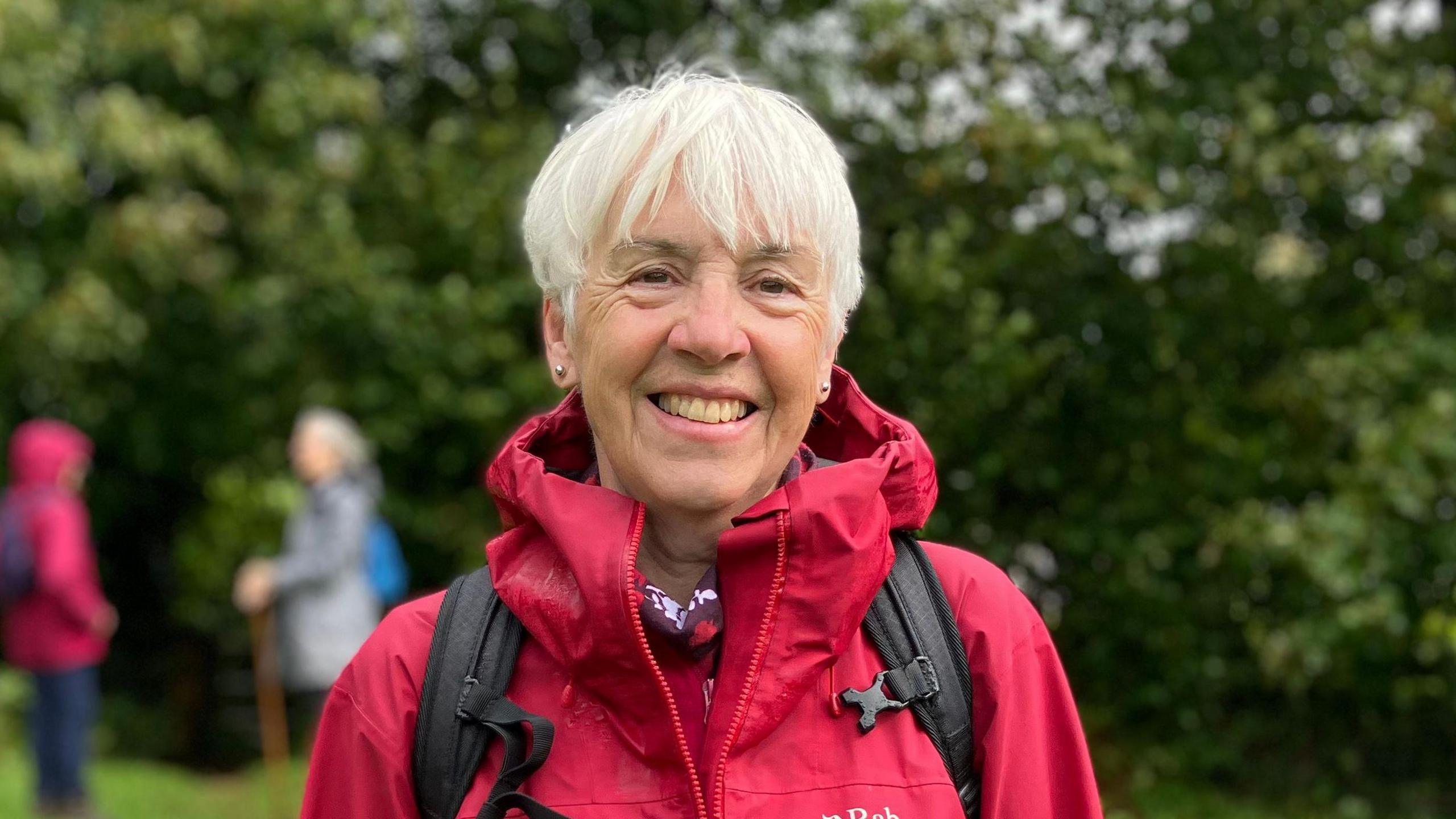 Woman with white hair and wearing a red coat and black rucksack. She