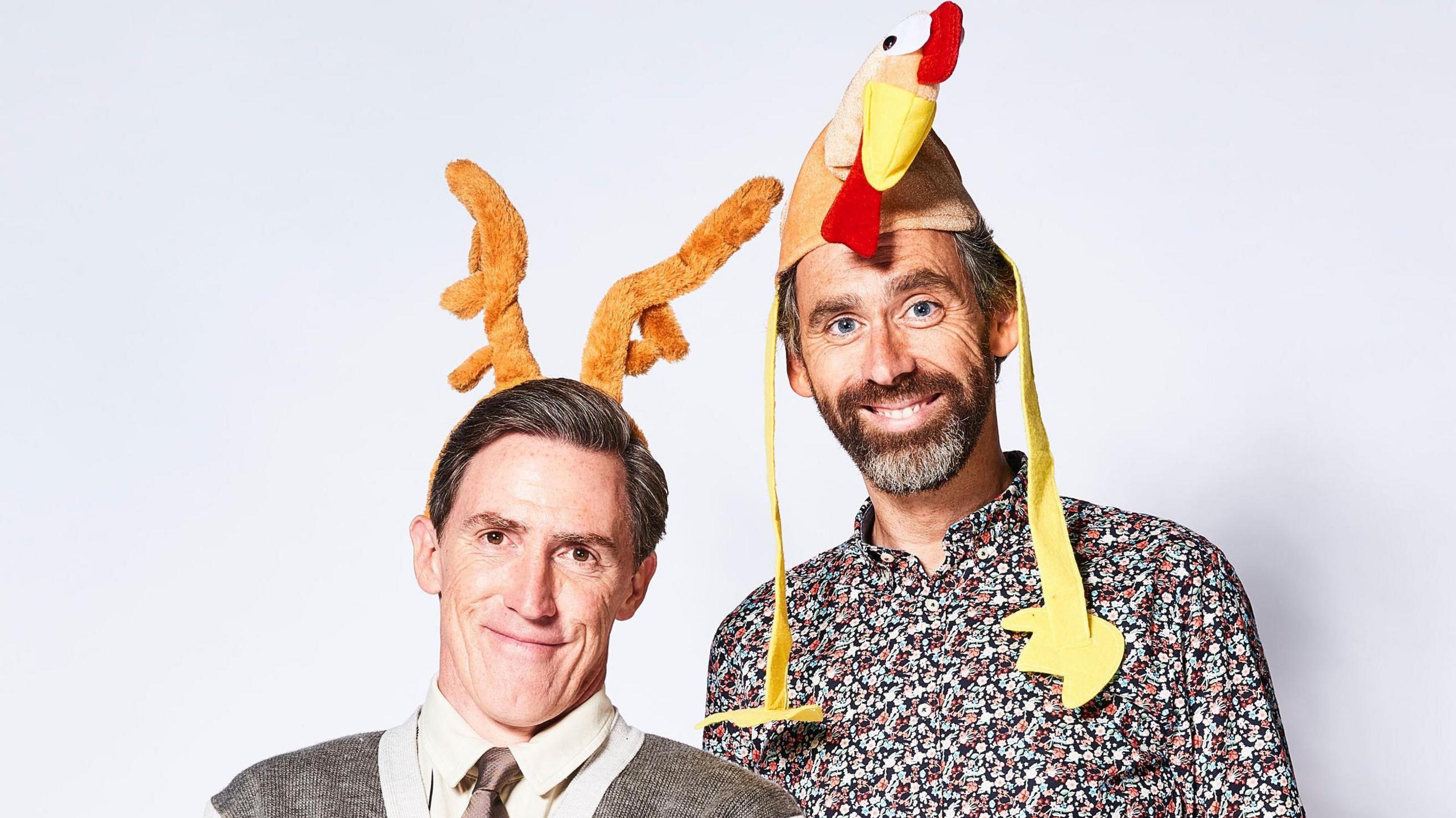 Rob Brydon and Robert Wilfort stood together against a white background wearing comical Christmas hats. Brydon has reindeer horns and Wilfort has a turkey.