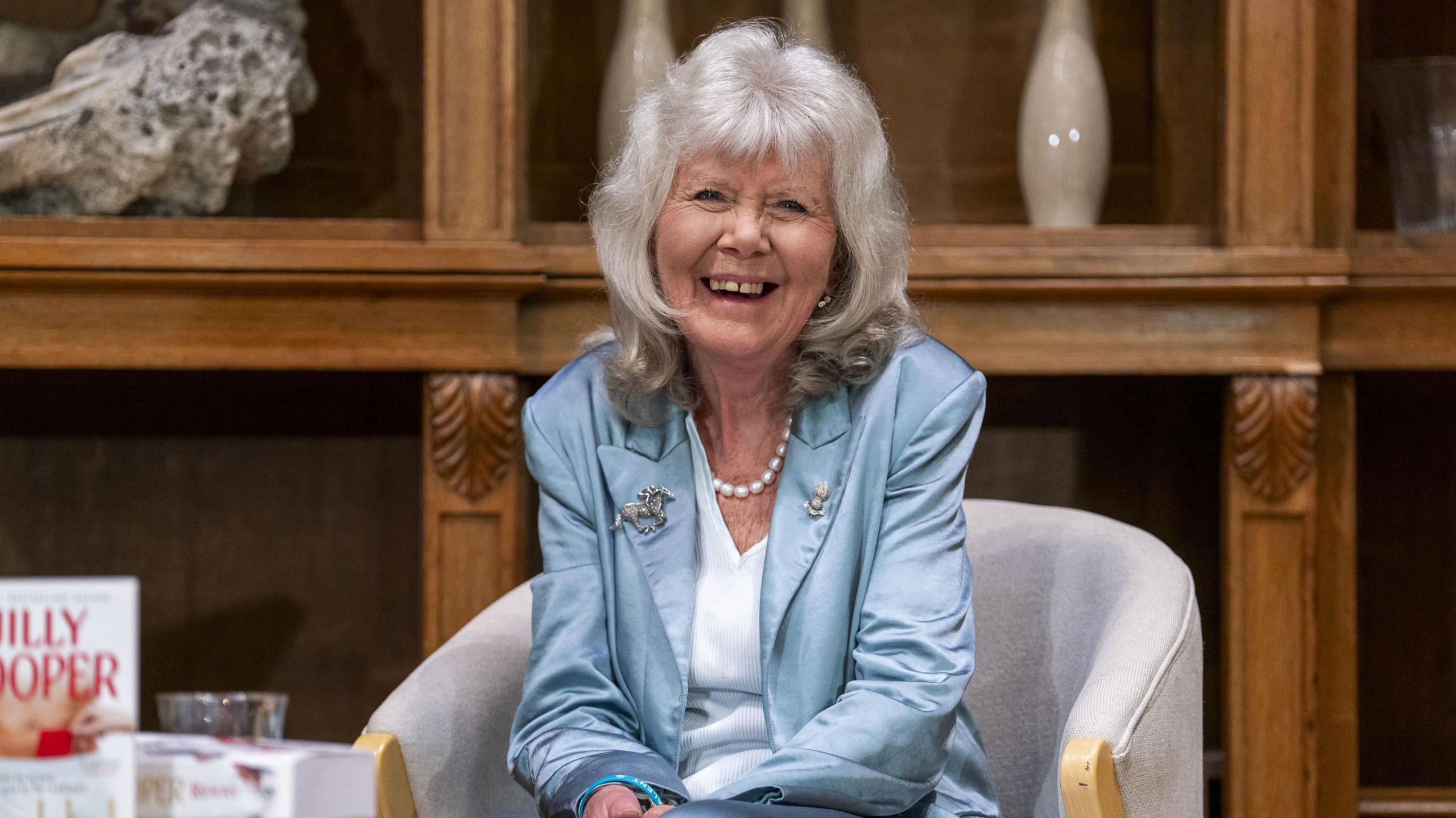 Dame Jilly Cooper laughs as she sits in a chair in front of a large, ornate, wooden shelving unit. She has grey hair to her shoulders and is wearing a blue satin suit jacket, a light blue V-neck top and a string of pearls around her neck. One of her books can be seen on a table in the foreground.