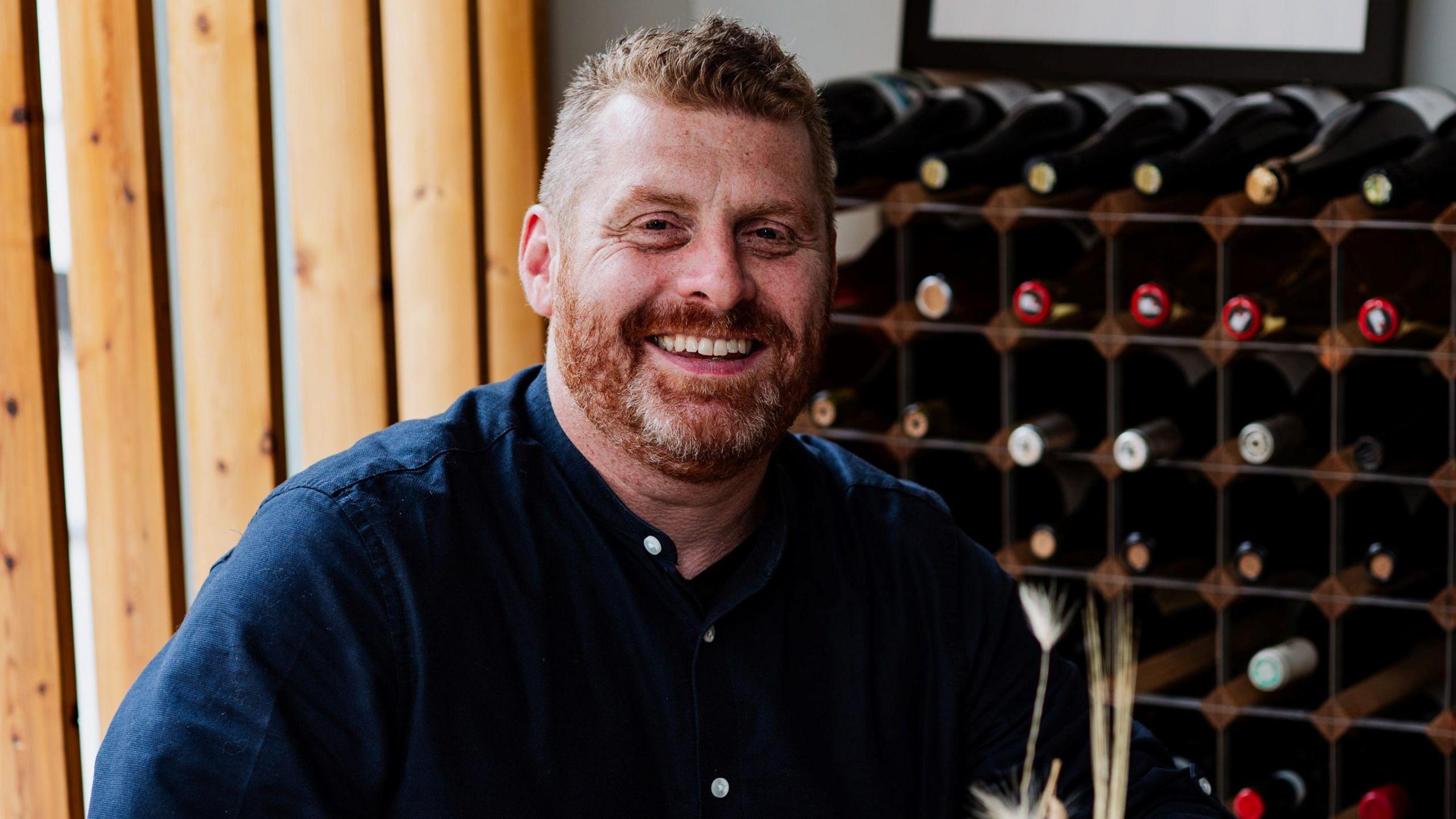 A man with short ginger hair and a ginger beard, and wearing a blue buttoned shirt, smiles as he sits in front of a large wine rack full of bottles of wine.