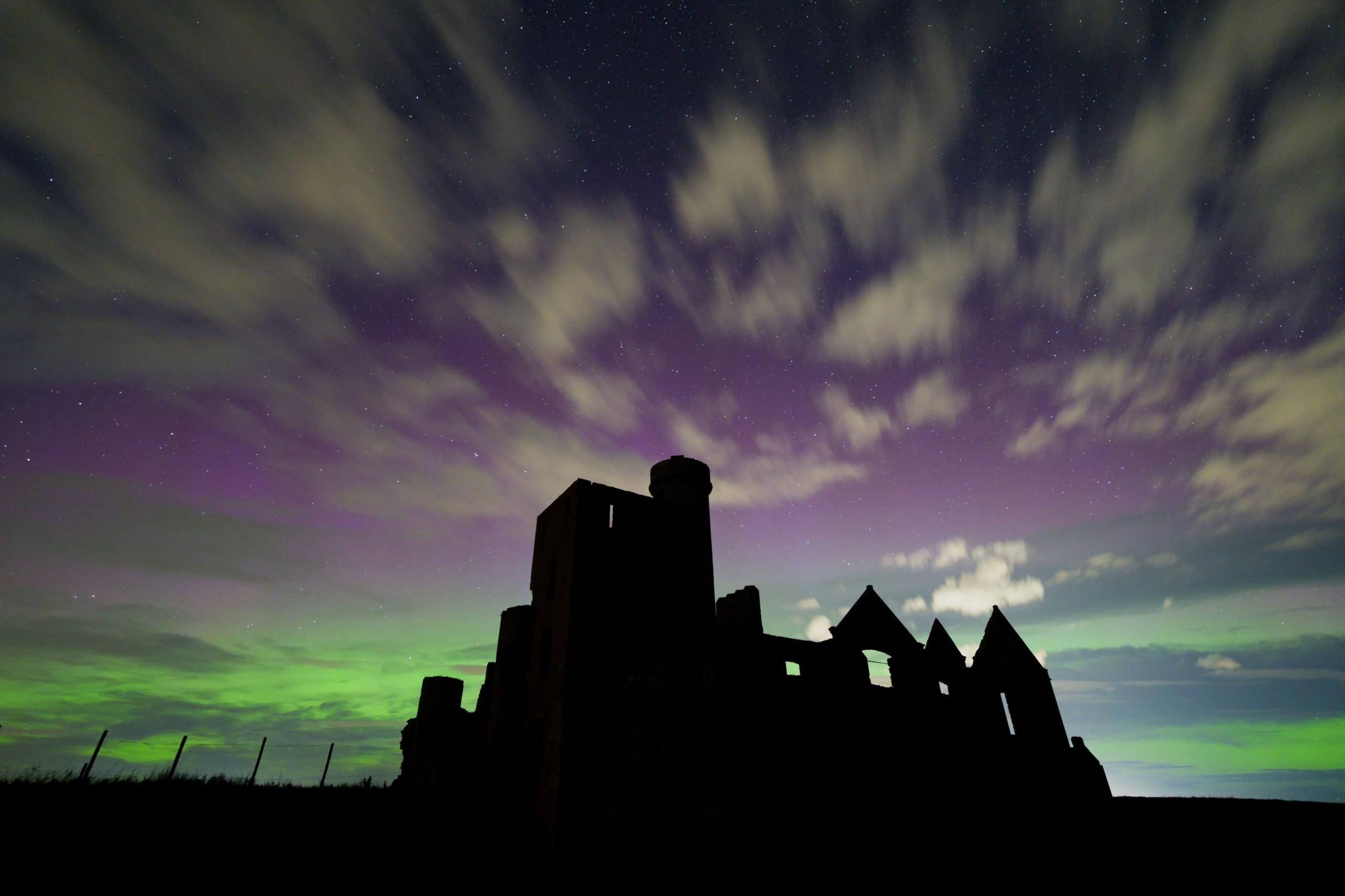 Slains Castle in Aberdeen is silhouetted against a colourful pink, green and blue display of the northern lights, with clouds and stars also visible.