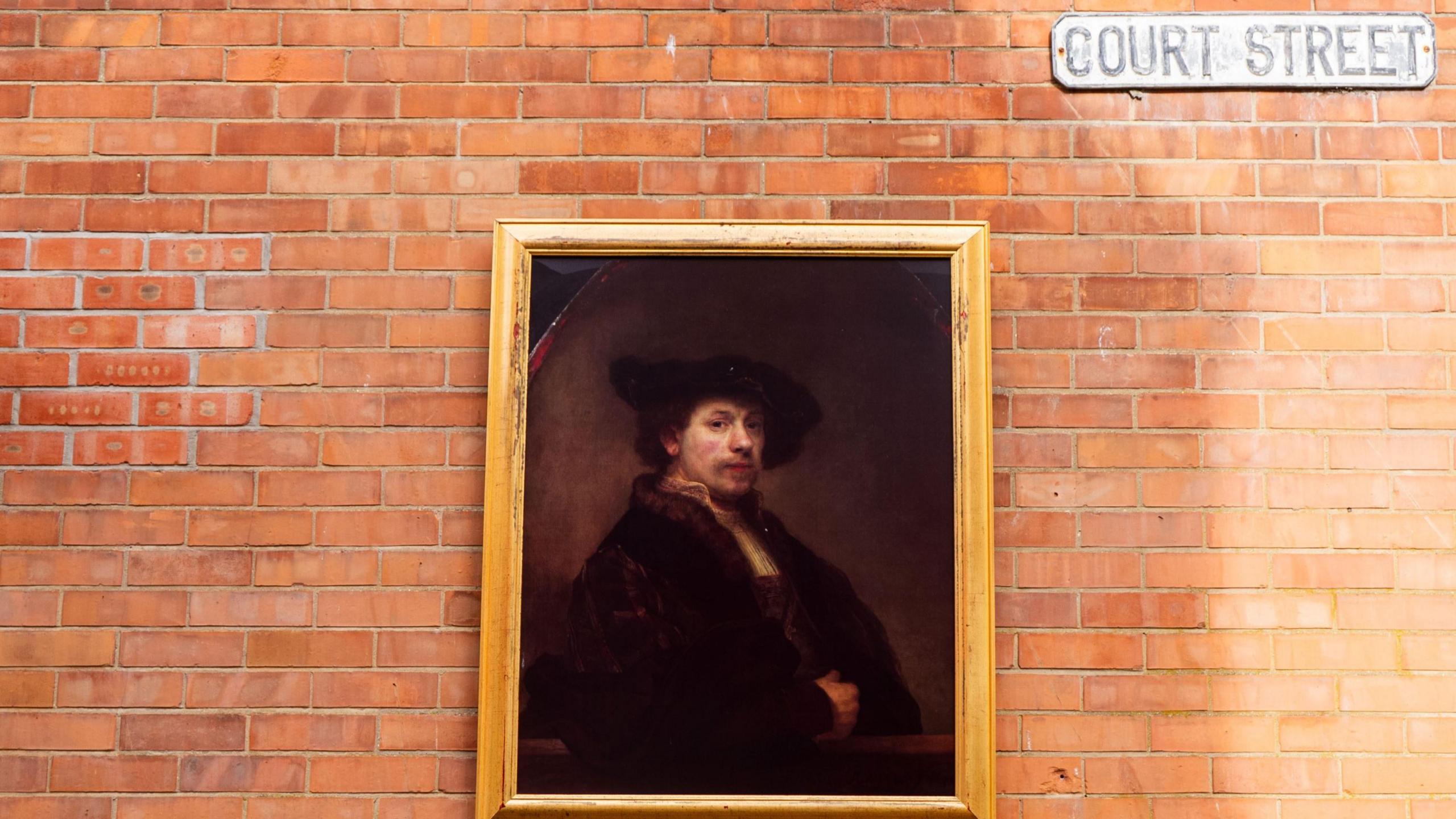 An image of the artwork Rembrant's Self Portrait at the Age of 34 hung on a brick wall with a street sign above which reads Court Street,
