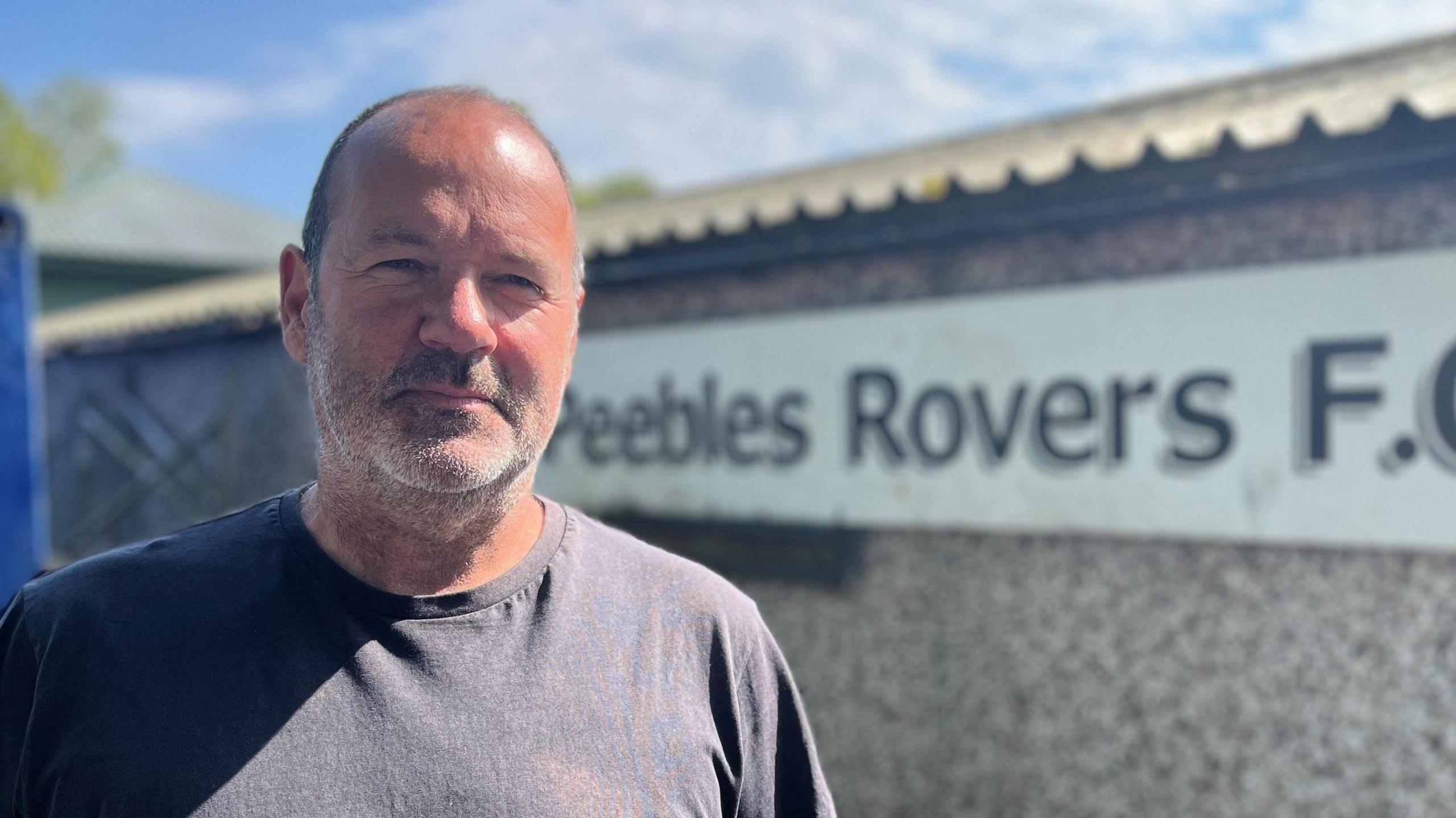 A bald-headed man with a short grey beard stands in front of a Peebles Rovers sign