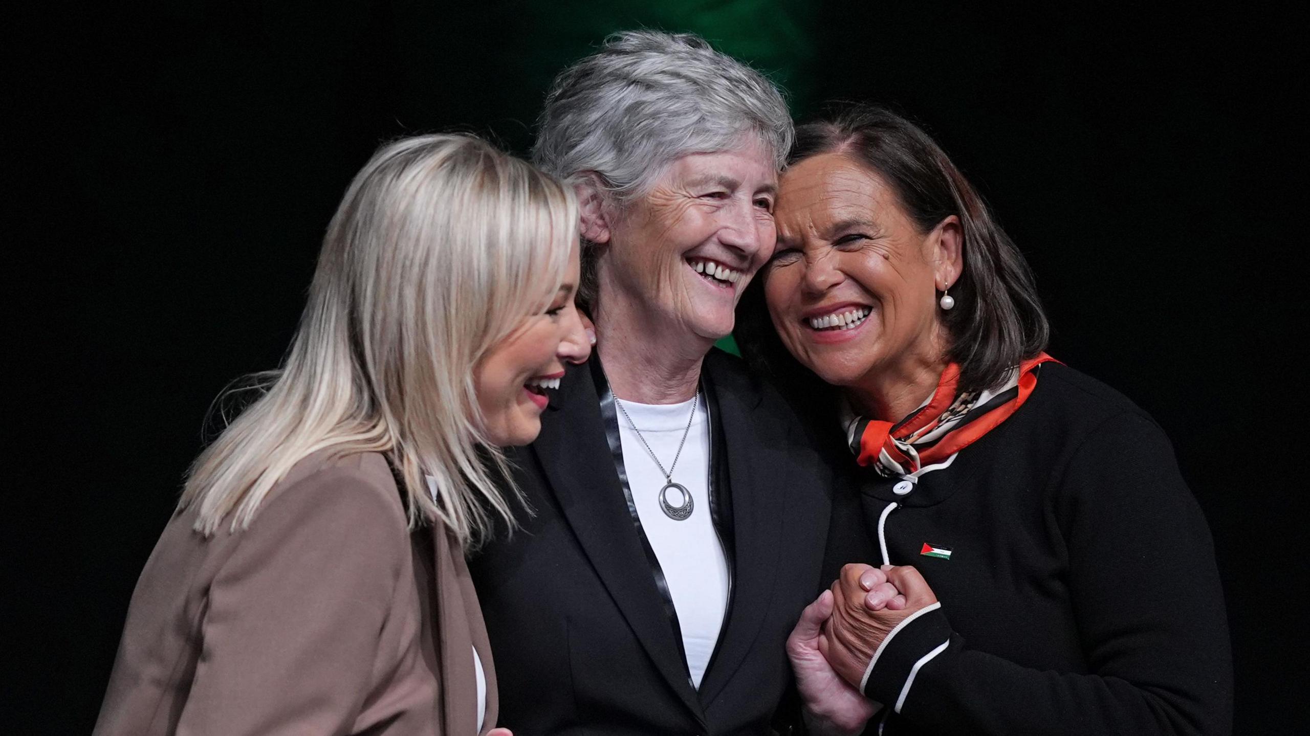 Sinn Féin Deputy leader Michelle O'Neill (left) and Sinn Féin Leader Mary Lou McDonald (right) hugging presidential candidate Catherine Connolly at a conference in Dublin.  All three women are smiling broadly.  McDonald is gripping Connolly's hand.