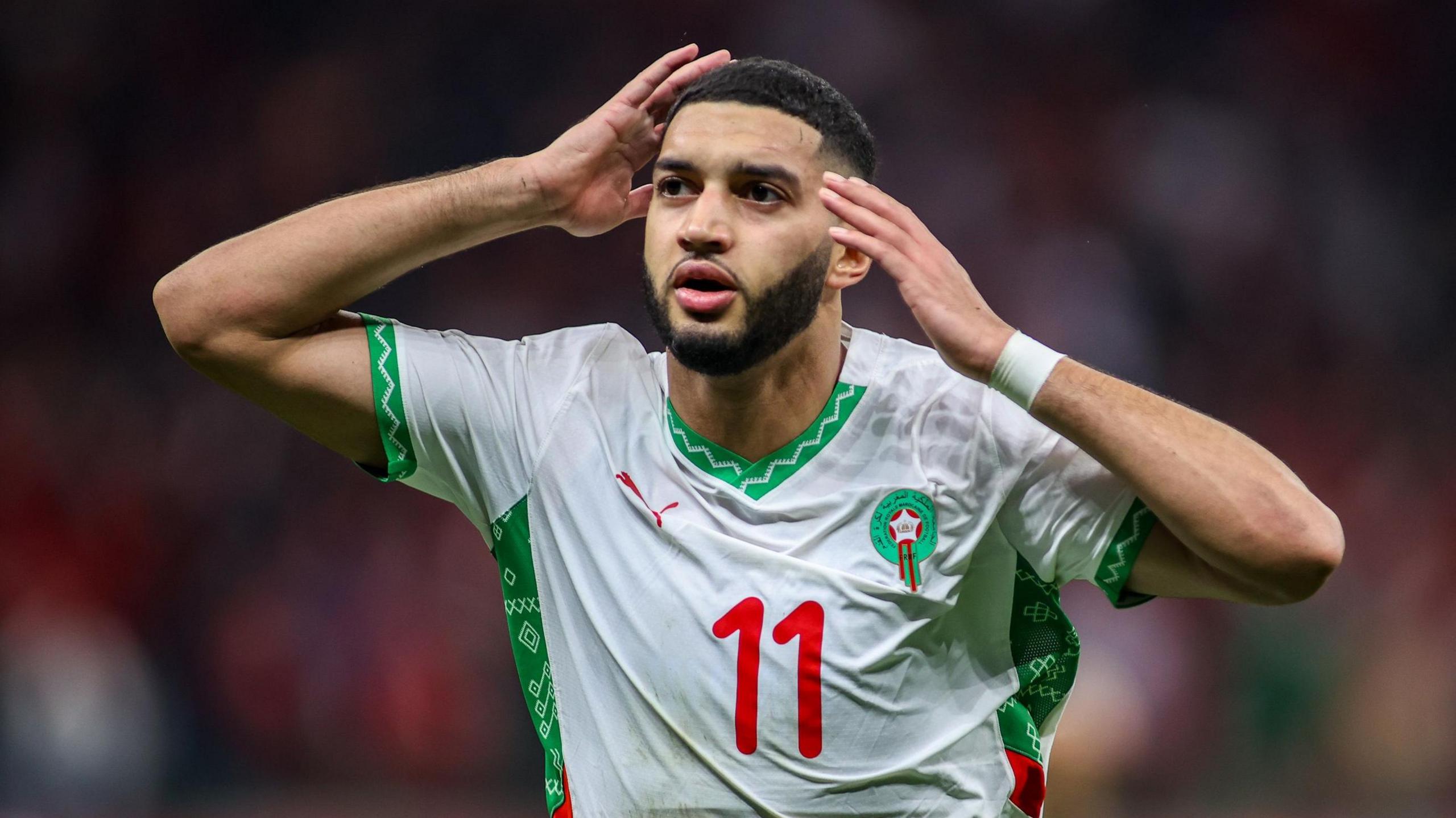 Ismael Saibari, wearing a white Morocco shirt with green trim and a red number 11 on the chest, puts his hands towards his head in celebration