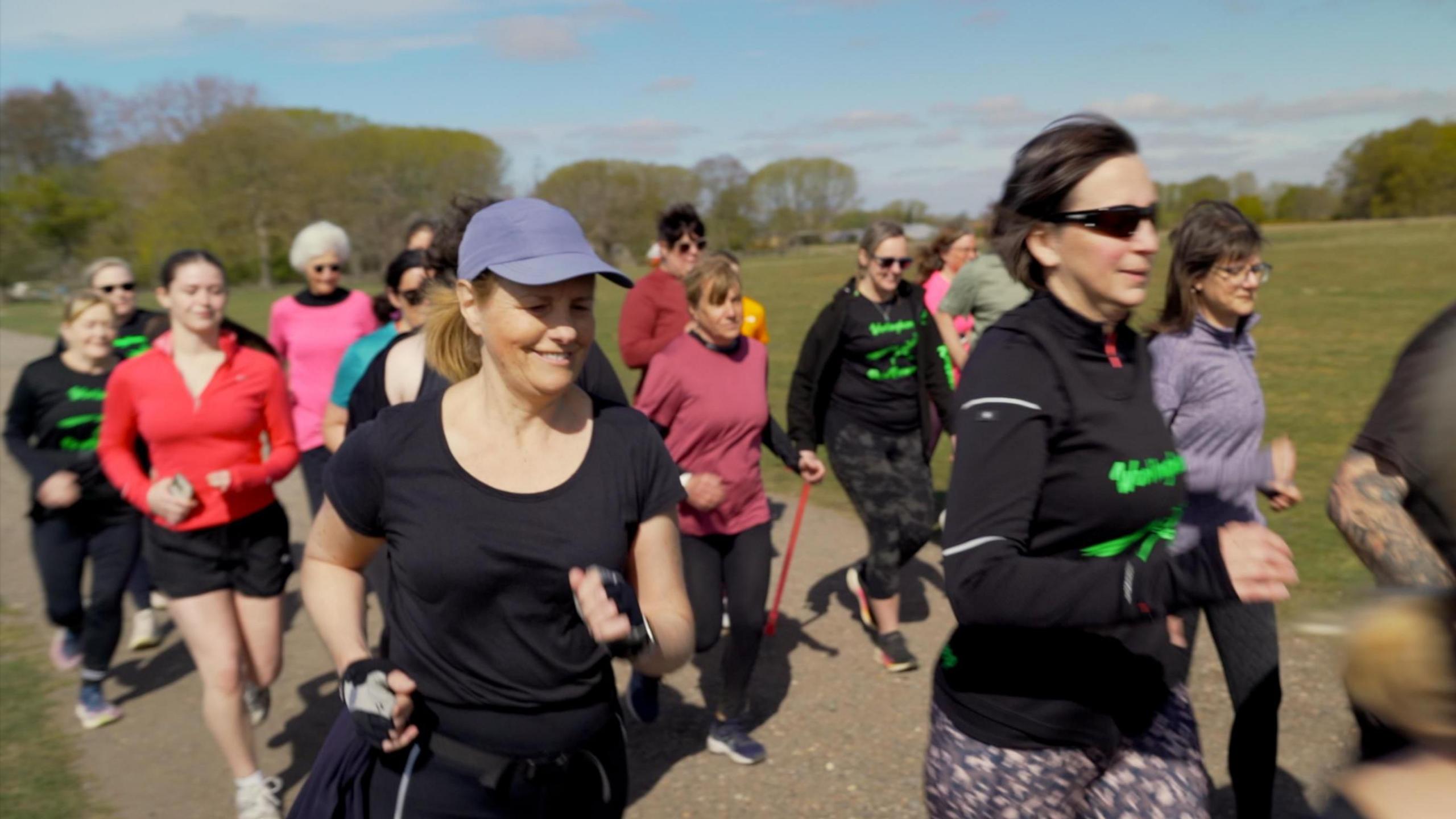 A group of runners zooming past the photographer. Upwards of 15 runners, many of which wearing the club's crest on their brightly coloured outfits. 

One of which is a lady, who is running whilst using a walking stick.