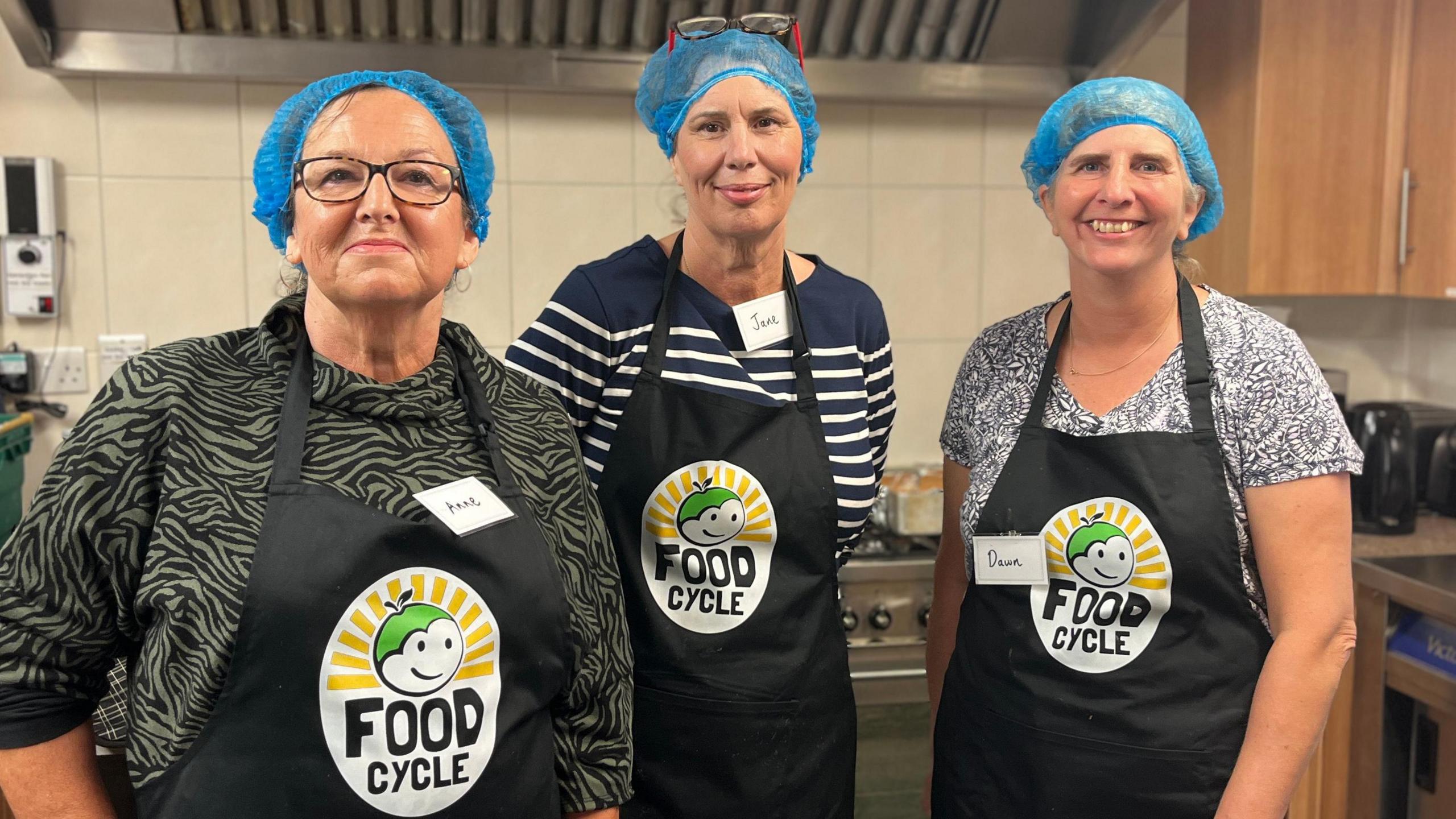 Ann Wyatt, Jane Hunt and Dawn Herbert stand in a kitchen wearing FoodCycle branded aprons and blue hairnets