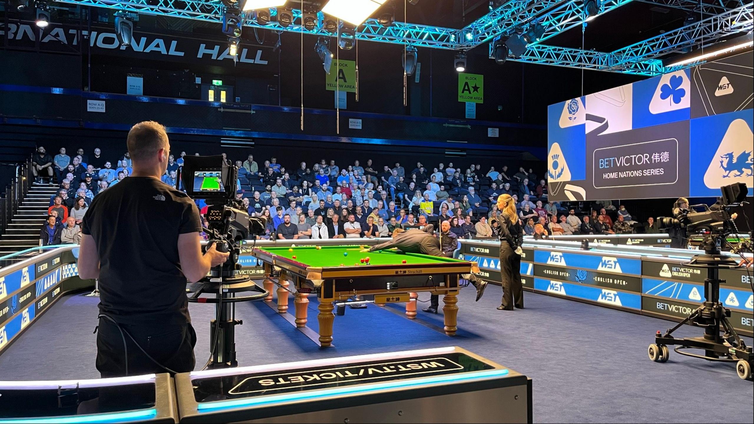 A snooker player is leaning on the table, taking aim for a shot. He is playing in a match at the Brentwood Centre. A referee wears all black as she stands behind him. In the foreground a camera operator films the action. In the background ar several rows of people in the crowd, all watching on.