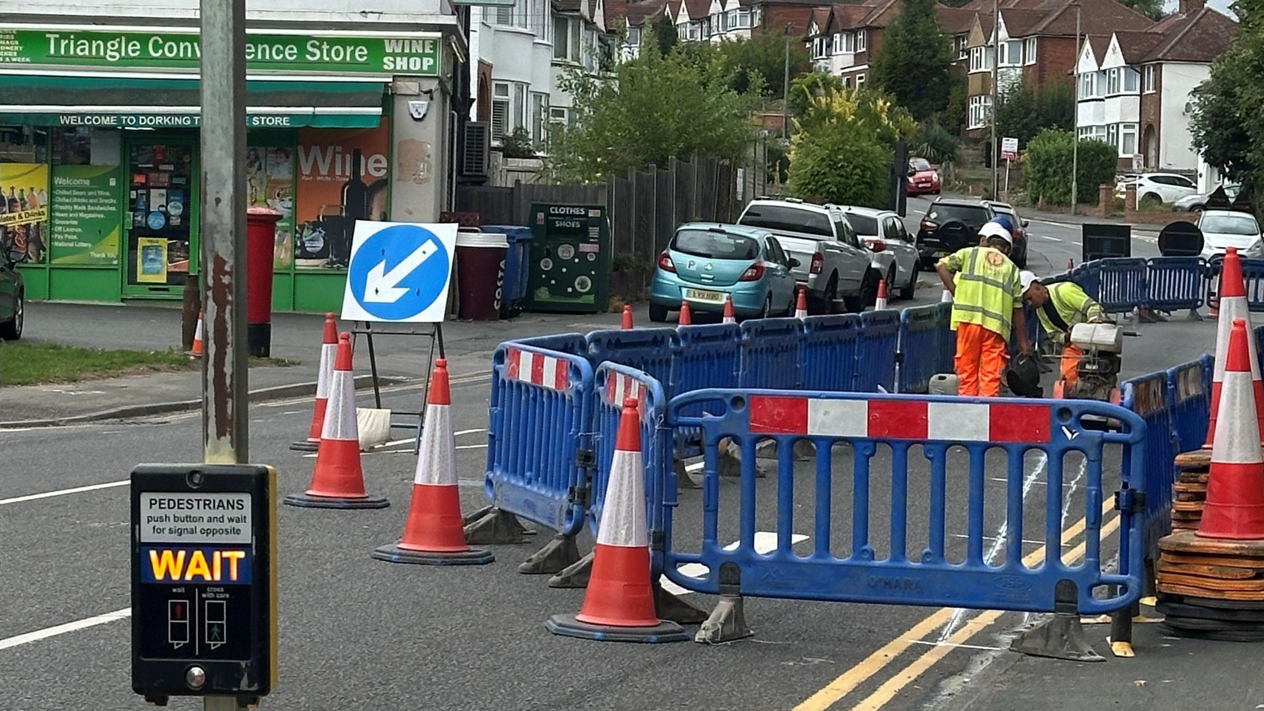 Builders in high vis jackets stand in a road surrounded by royal blue barriers and red and white traffic cones as they mark out an area of widened pavement using white paint. A convenience store and a number of houses and cars can be seen in the background.
