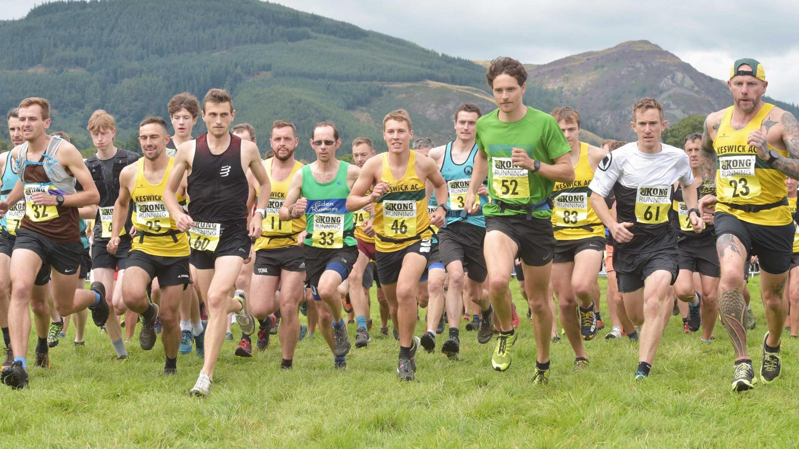 A group of young men wearing numbers running uphill with the hills of the Lake District behind them.