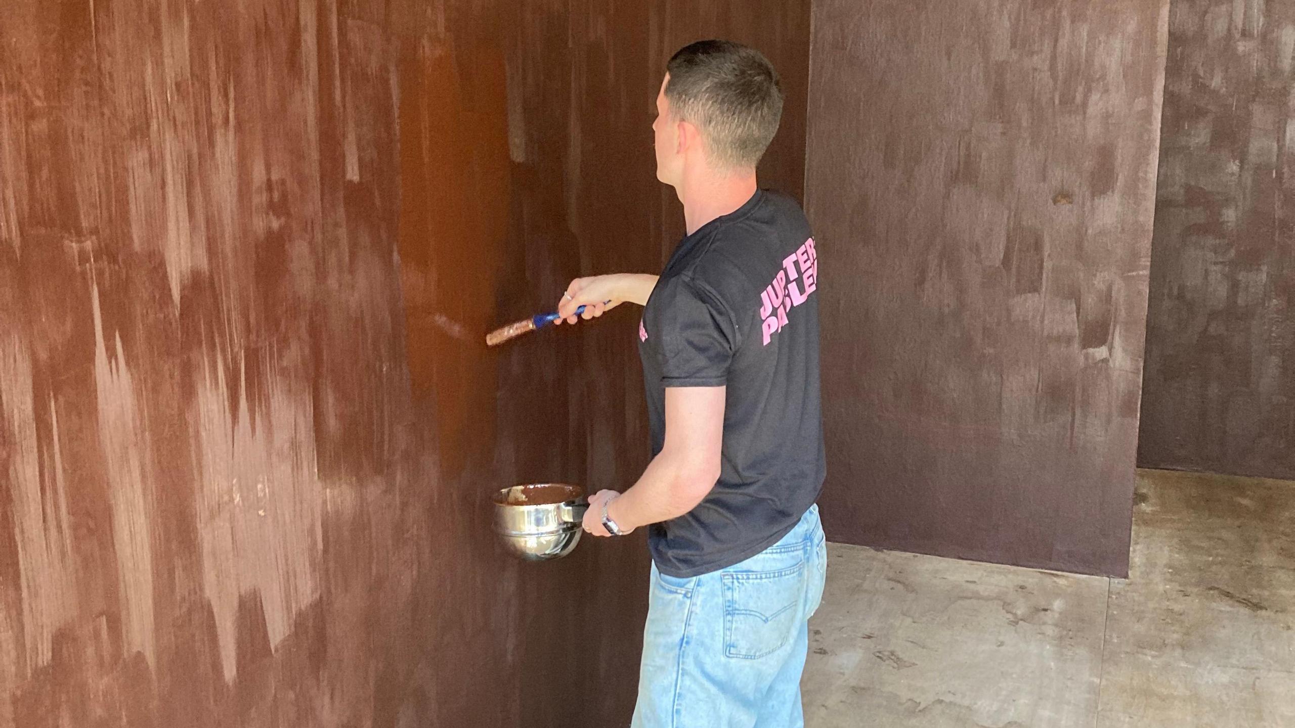 A man with a bowl of chocolate paint brushes the chocolate onto the wall. He is wearing a black shirt with pink lettering.