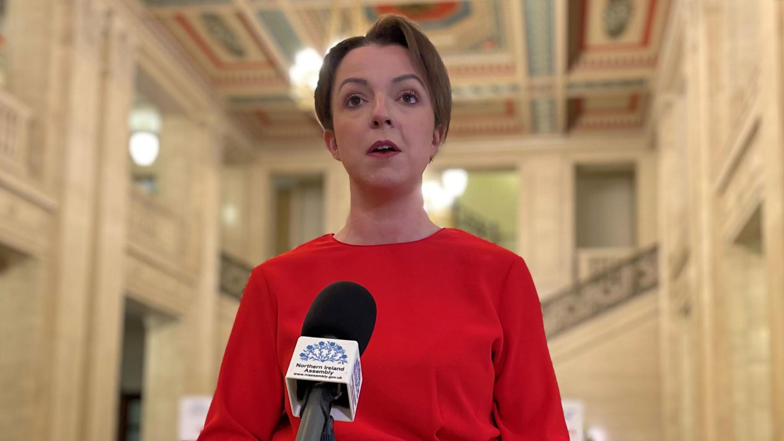 Kimmins is standing in front of a mic in the hall at Stormont. She's wearing red. Marble steps are behind her.