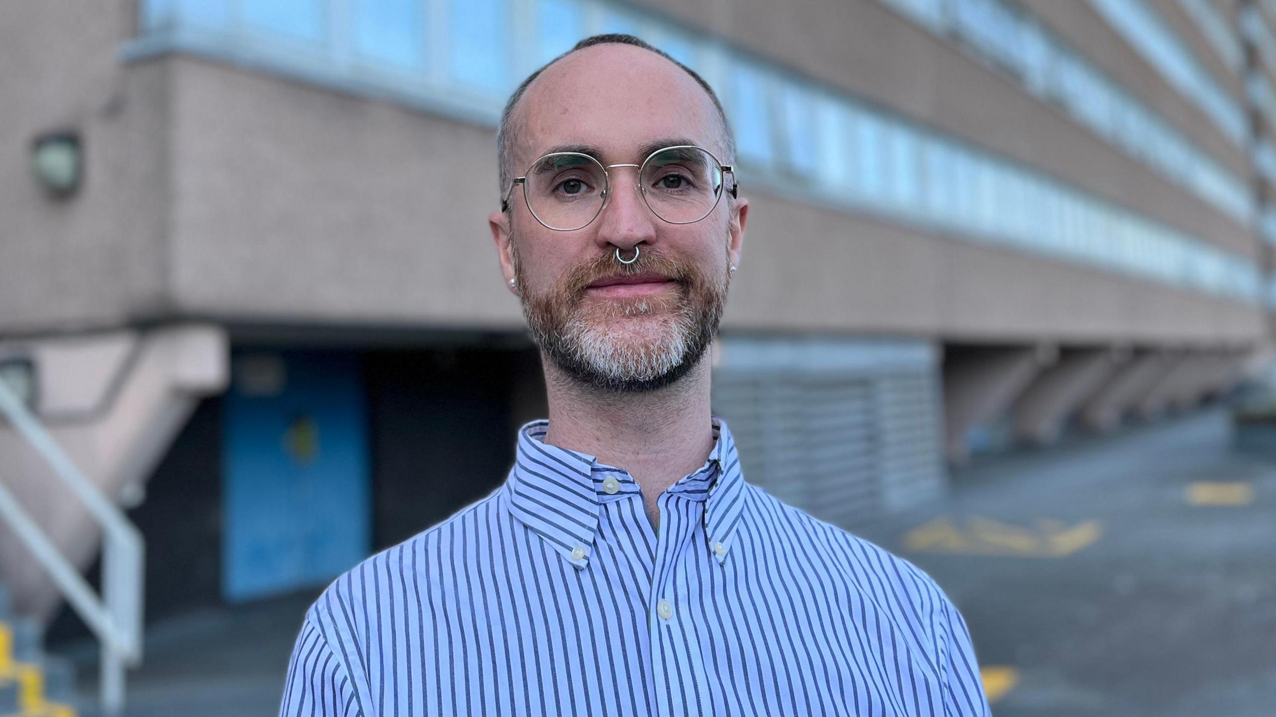 Liam McClelland a 37-year-old man of a slim build, wearing a blue pinstripe shirt. He has a short greying beard a septum piercing and is wearing glasses with a metal frame.