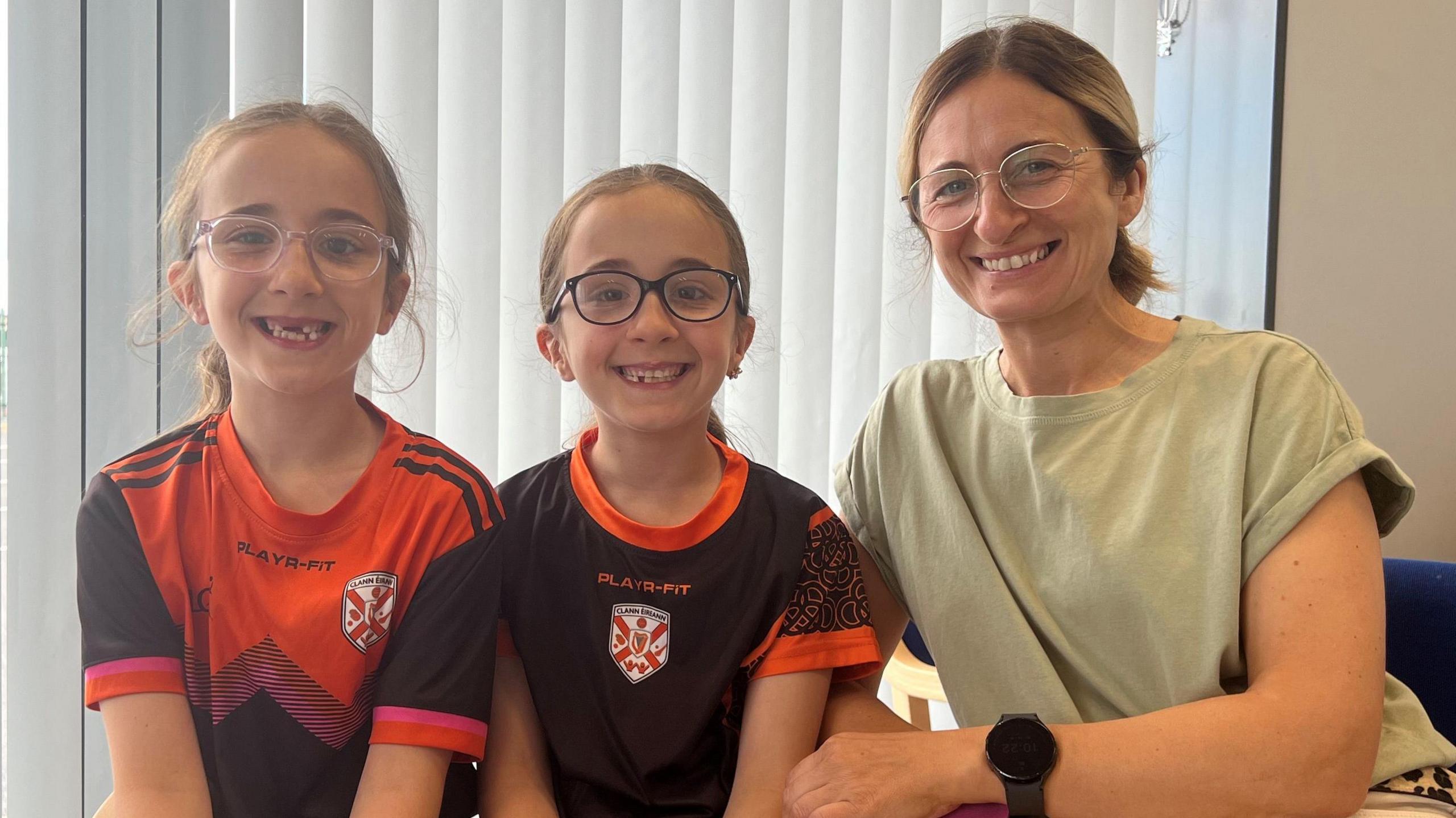 Natalie Geoghegan has dark blonde hair pulled back into a ponytail. She is wearing a light green short-sleeved top and white trousers and is sitting with two girls. She and the girls are all wearing glasses. The two girls are wearing sport tops which are black and red with a white and red logo in the middle.