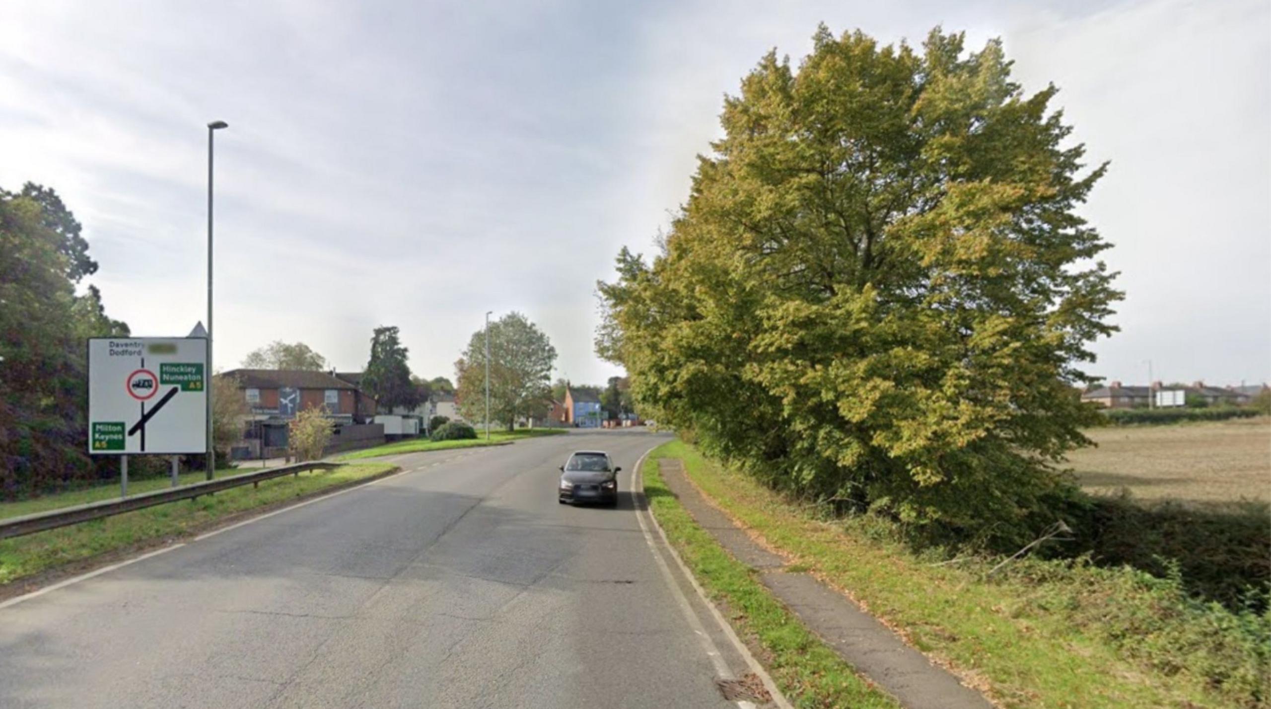 The road at the top of a hill. There is one car travelling towards the camera. There is a large square, white roadsign to the left, showing the A5 crossing the road ahead. There is a pub/restaurant beyond the sign, and a large tree to the right of the road and pavement.