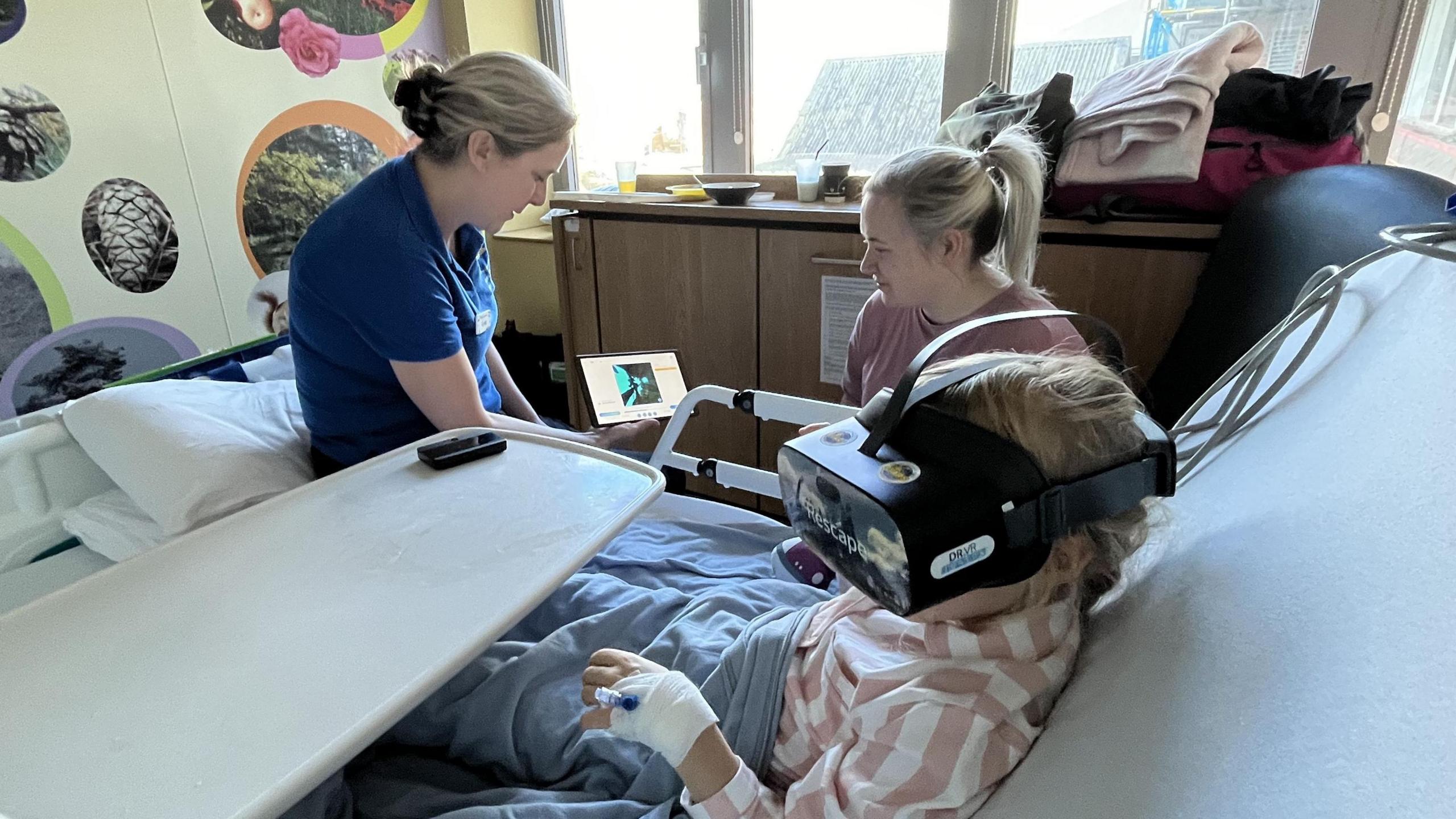 Play Assistant, Emma Lewis, shows Charlotte Cummings what her daughter, Freya, is seeing in the headset as they sit next to Freya's hospital bed