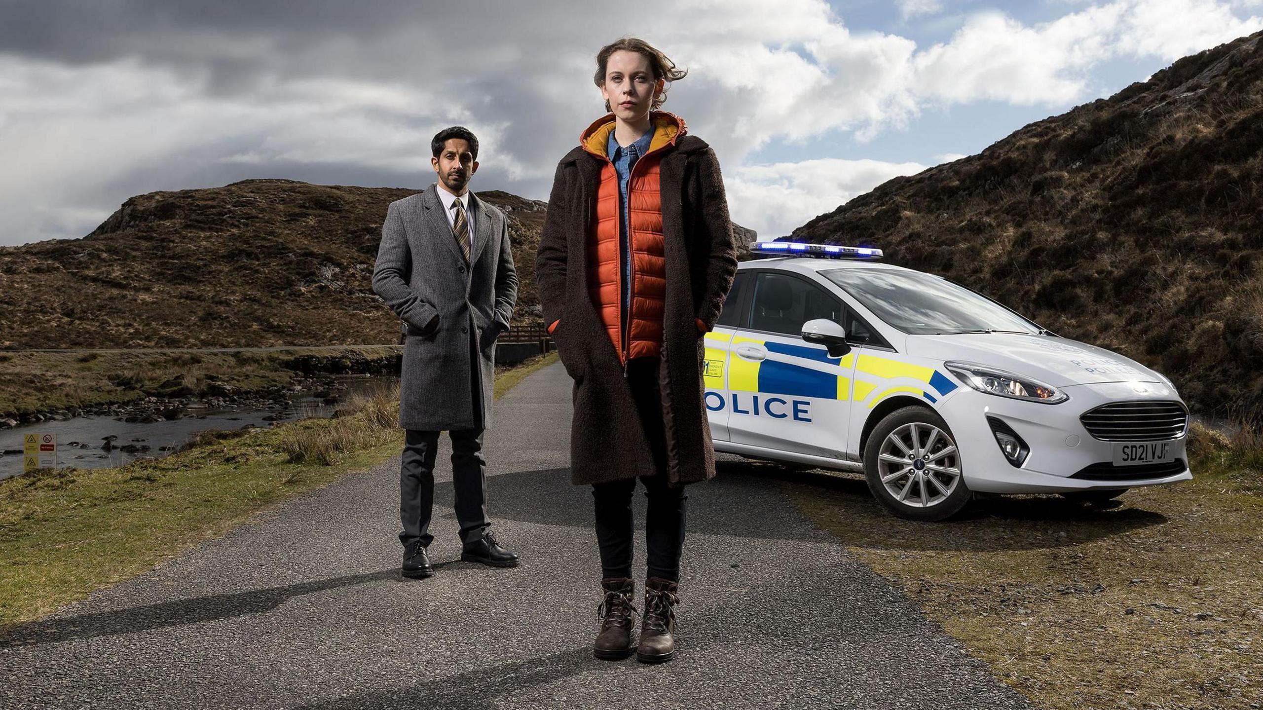 Radia, dressed in long grey coat, and Groundsell, wearing a woollen coat over an orange jacket, are standing in front of a police car on a single-track island road. 