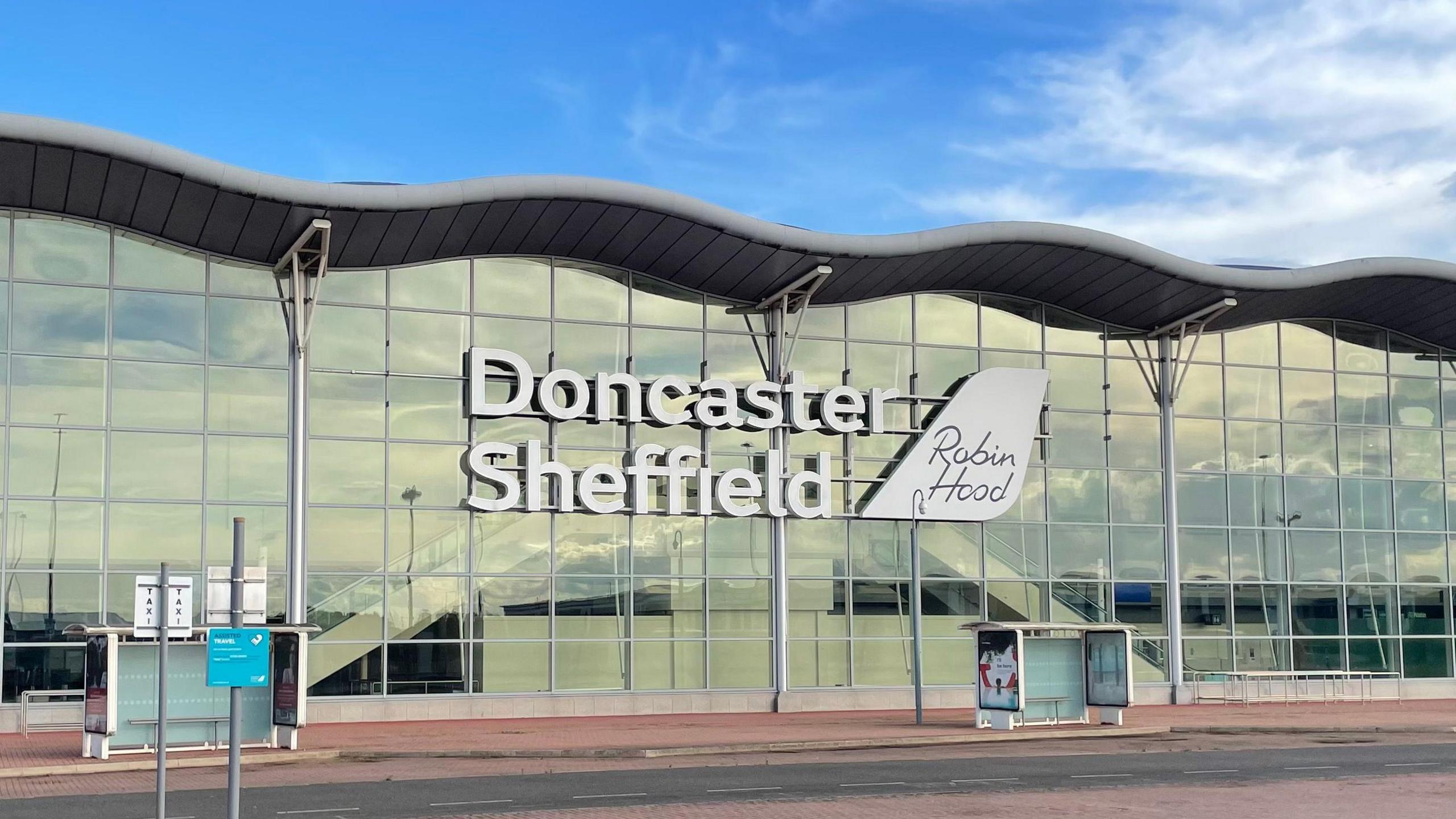 Against a backdrop of a blue sky, a wide glass fronted building with a wavy roof is at the side of a road. There are two bus stops spaced out in front of the building. Large white letters on the front of the building read Doncaster Sheffield. A white sign to the right of them reads Robin Hood.