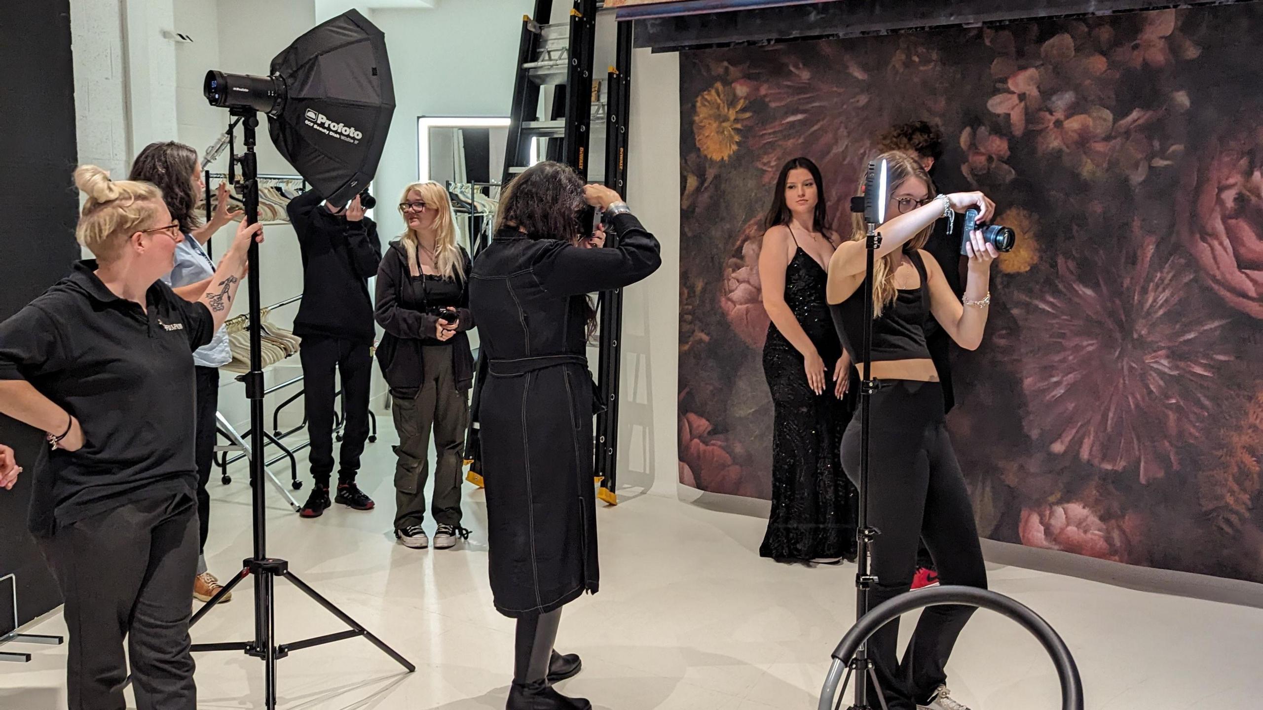 A photo taken of a photo shoot at Fujifilm Home of Production, showing three people holding cameras, a model posing for an image and other people standing around helping, all wearing black clothing