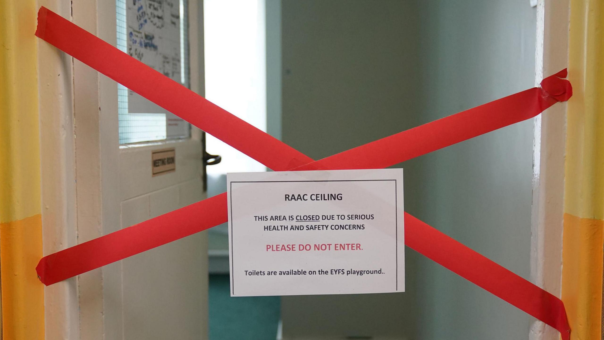 A classroom with an open door and red tape in a cross over the doorway warning about a Raac ceiling and saying "please do not enter".