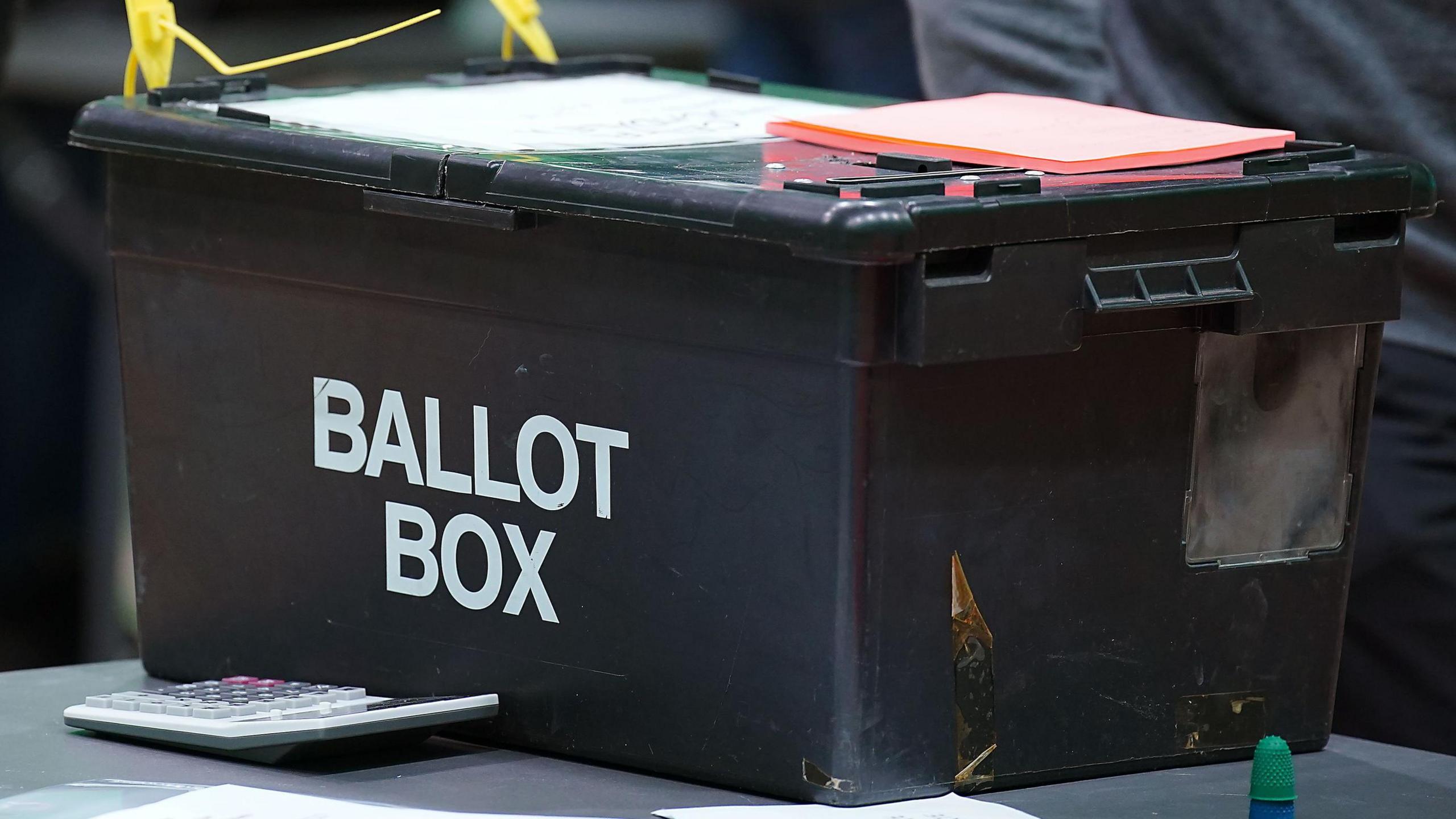 A black ballot box at a vote count. The words ballot box are written in white block capitals on the side of the box. A large calculator is next to the box, which is on a grey table. Pieces of orange paper are on top of the box.