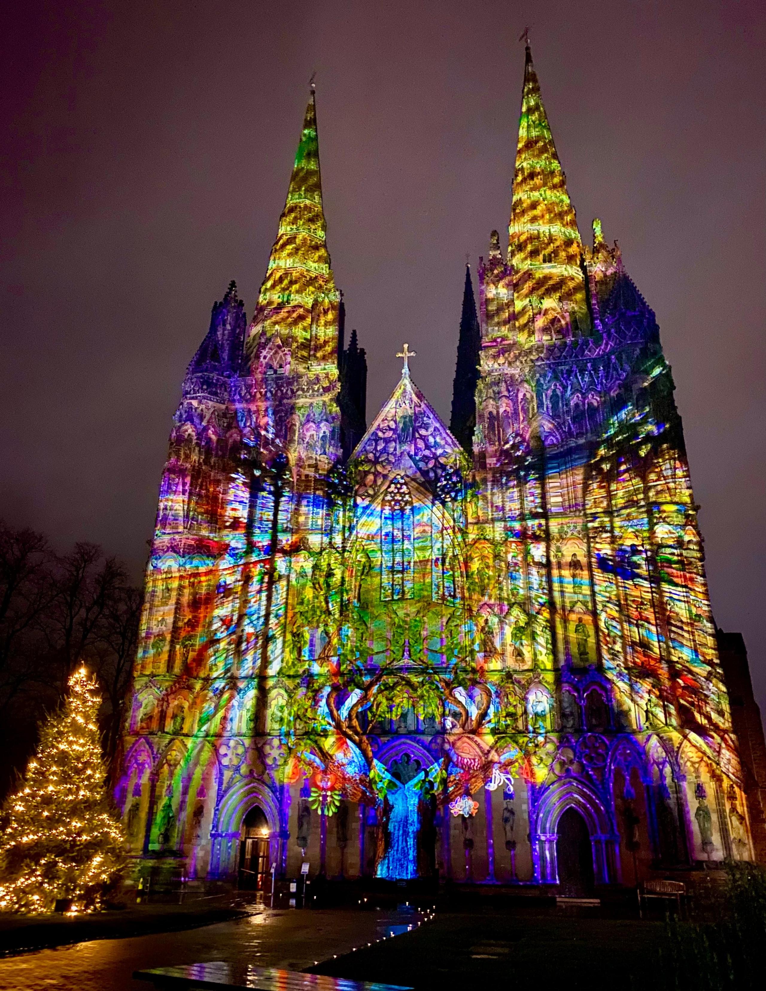 The front of a cathedral at night with a bright, abstract, multi-coloured image projected on the front. A Christmas tree stands to the left.  