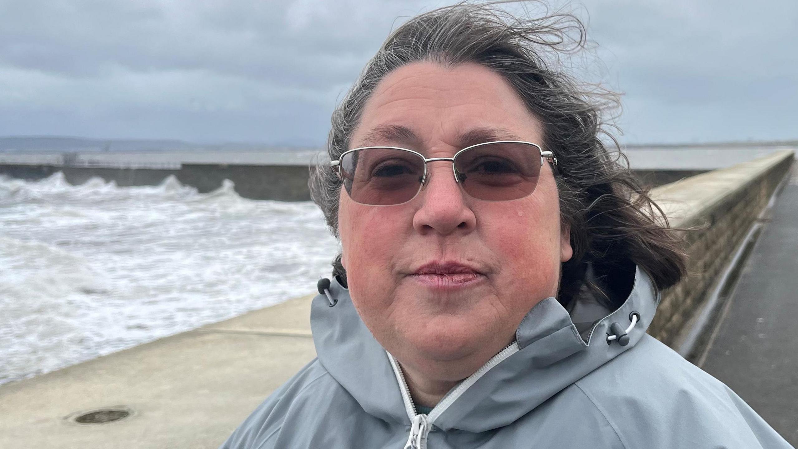 Dawn Lord has light brown sunglasses on with her hair being blown back in the wind. She is wearing a light grey coat with the sea splashing against a sea wall in the background.