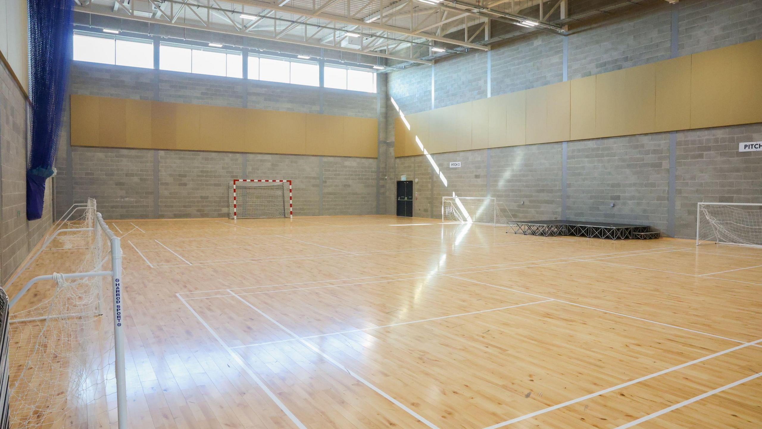 The inside of an empty sports hall. There is a wooden floor and grey brick walls. 