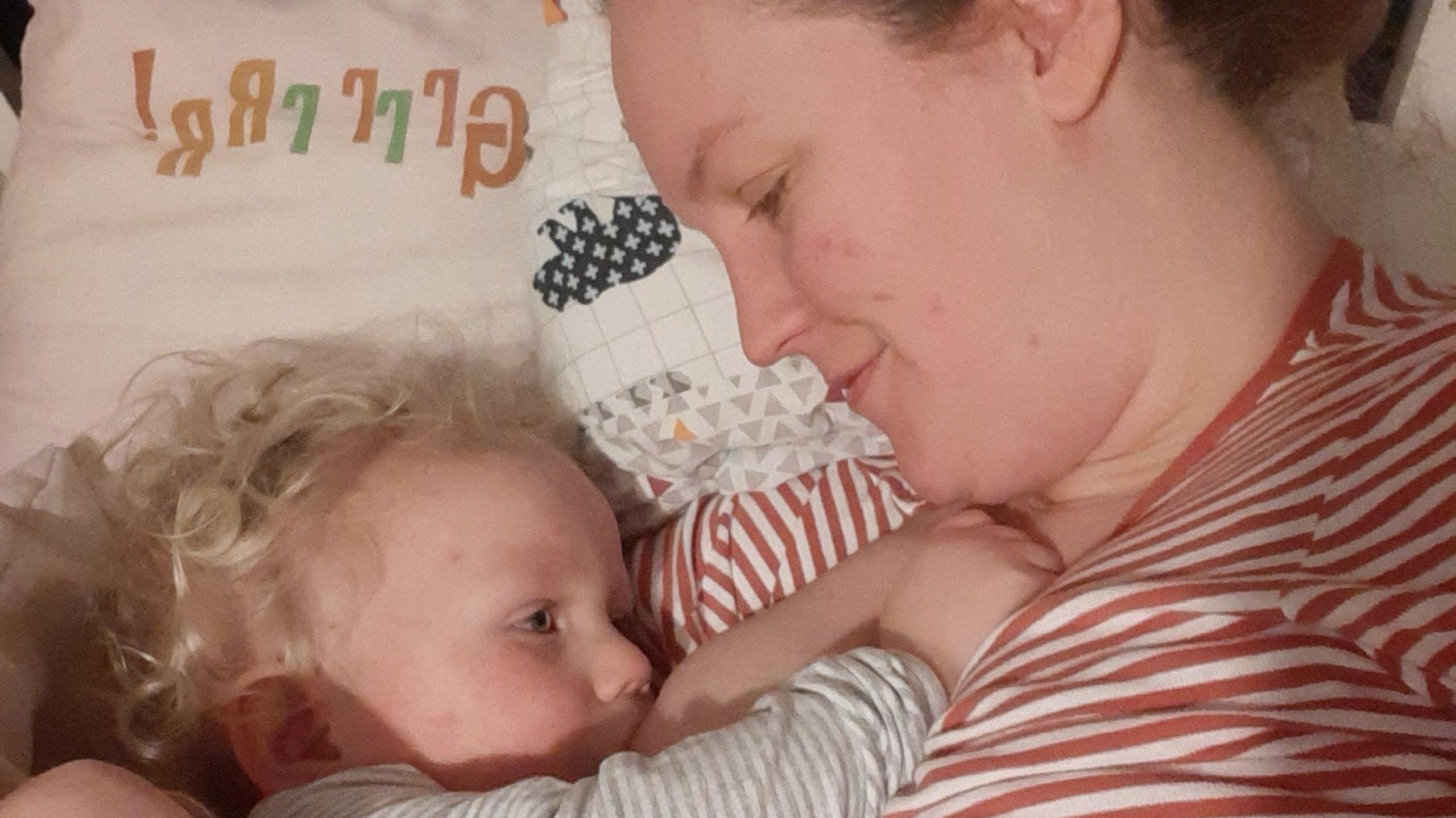 Josie is pictured smiling down at her little boy as he breastfeeds. The mother and son are lying together on a bed, she has her hair up and is wearing a red-and-white striped top; her baby son has blond hair and wears a grey-and-white top.