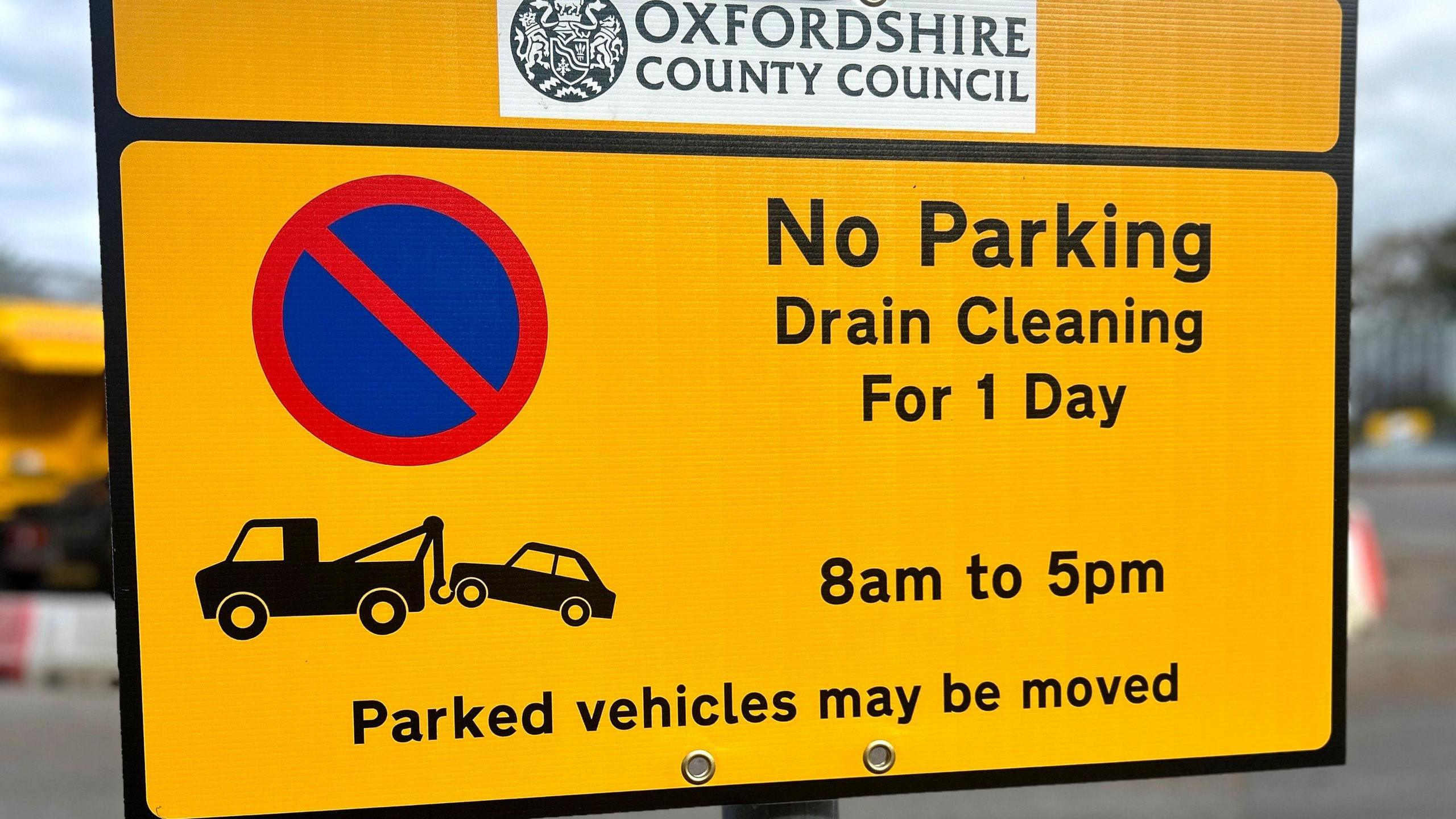 A yellow street No Parking sign by Oxfordshire County Council. It says Drain Cleaning for 1 Day 8am to 5pm and that Parked vehicles may be removed.