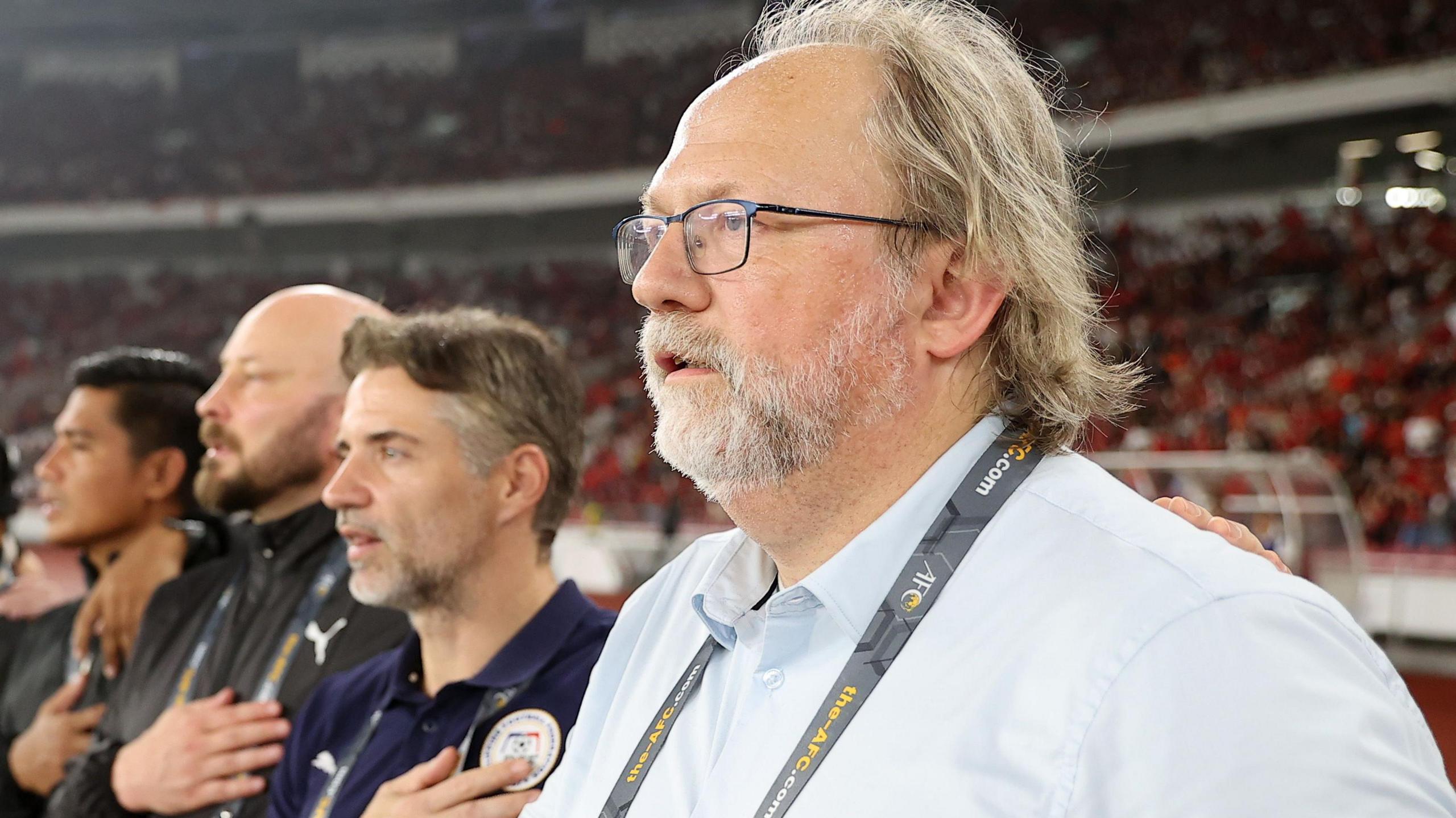 Tom Saintfiet is seen from chest up, wearing white shirt, glasses and beard, standing on the touchline at a football stadium as three men are seen in the background alongside him with hand on heart while respecting a national anthem.