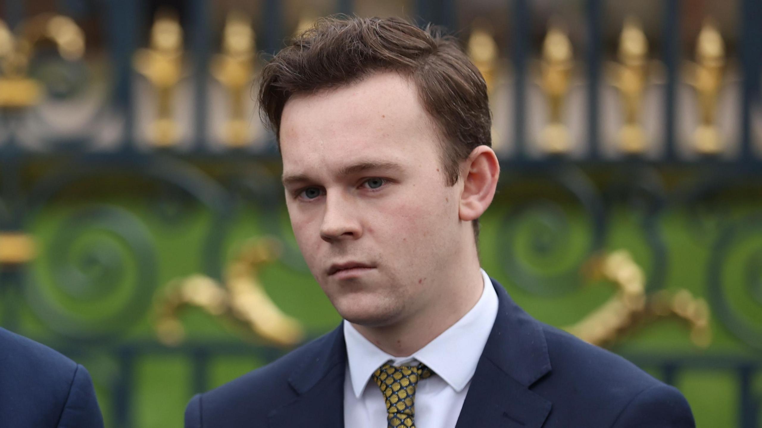 Eoin Tennyson - a man with short, brown hair is standing in front of an black and gold iron gate. He is wearing a dark suit jacket, a white collared shirt and a pattern yellow tie.