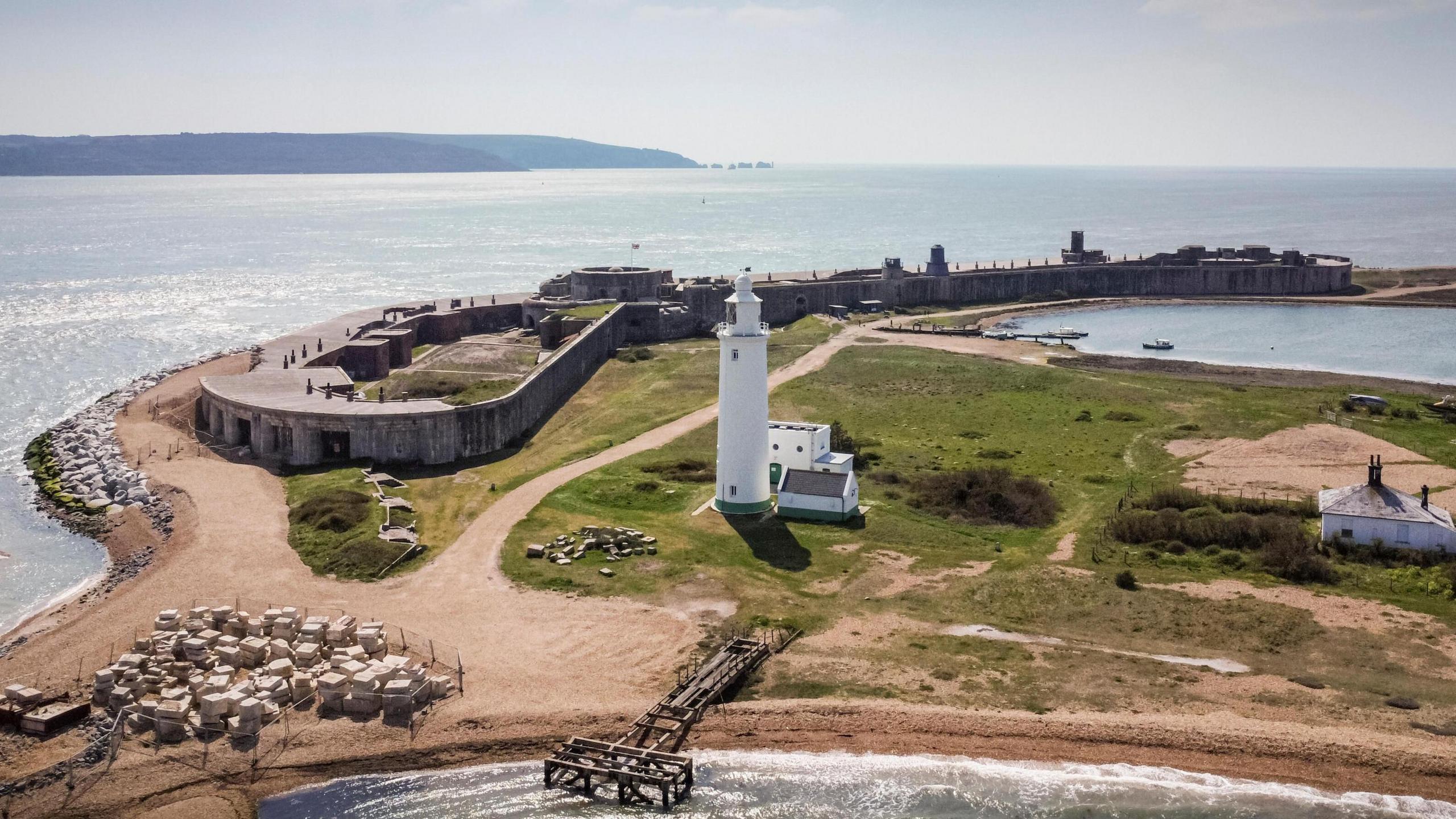 Hurst castle, with its low ceilings and perimeter spanning the tip of the shingle bank, and a white lighthouse, pictured at the tip of Hurst Spit in the foreground against the sea in the background. 