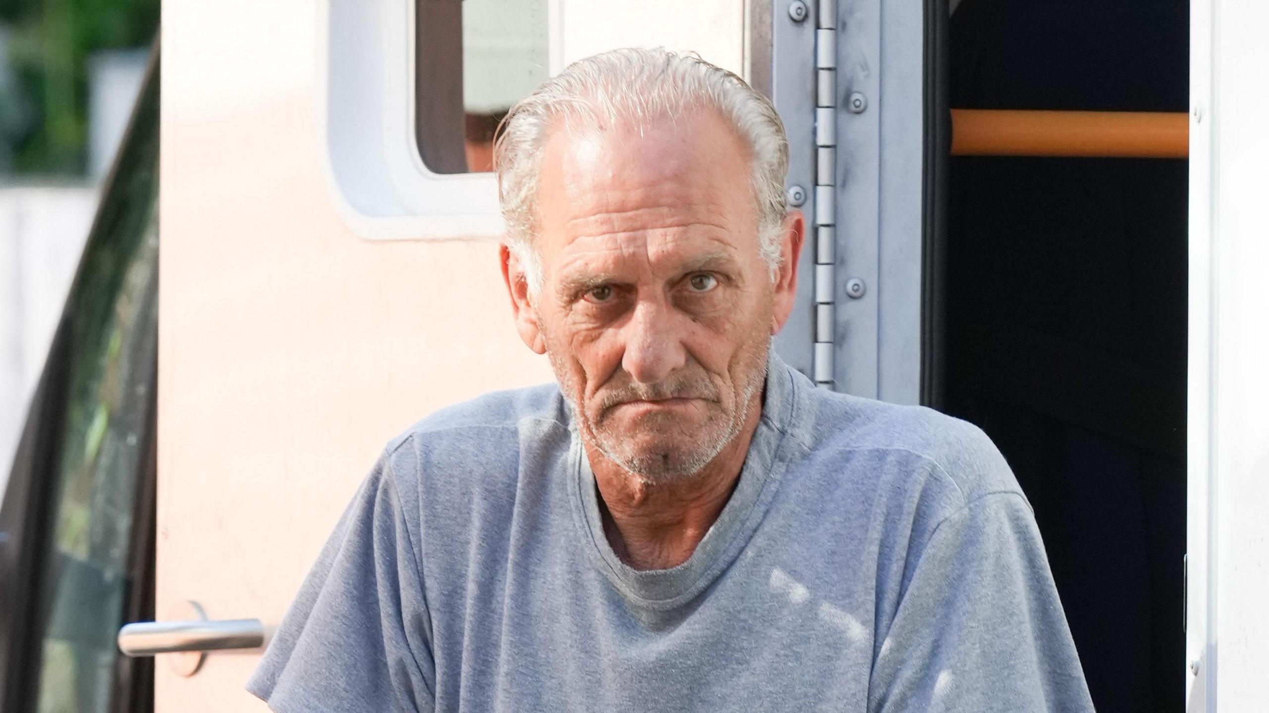 Derek Martin steps out of a prison van as he arrives at court. He has grey hair combed back and a grey stuble beard. He wears a grey roundneck t-shirt.
