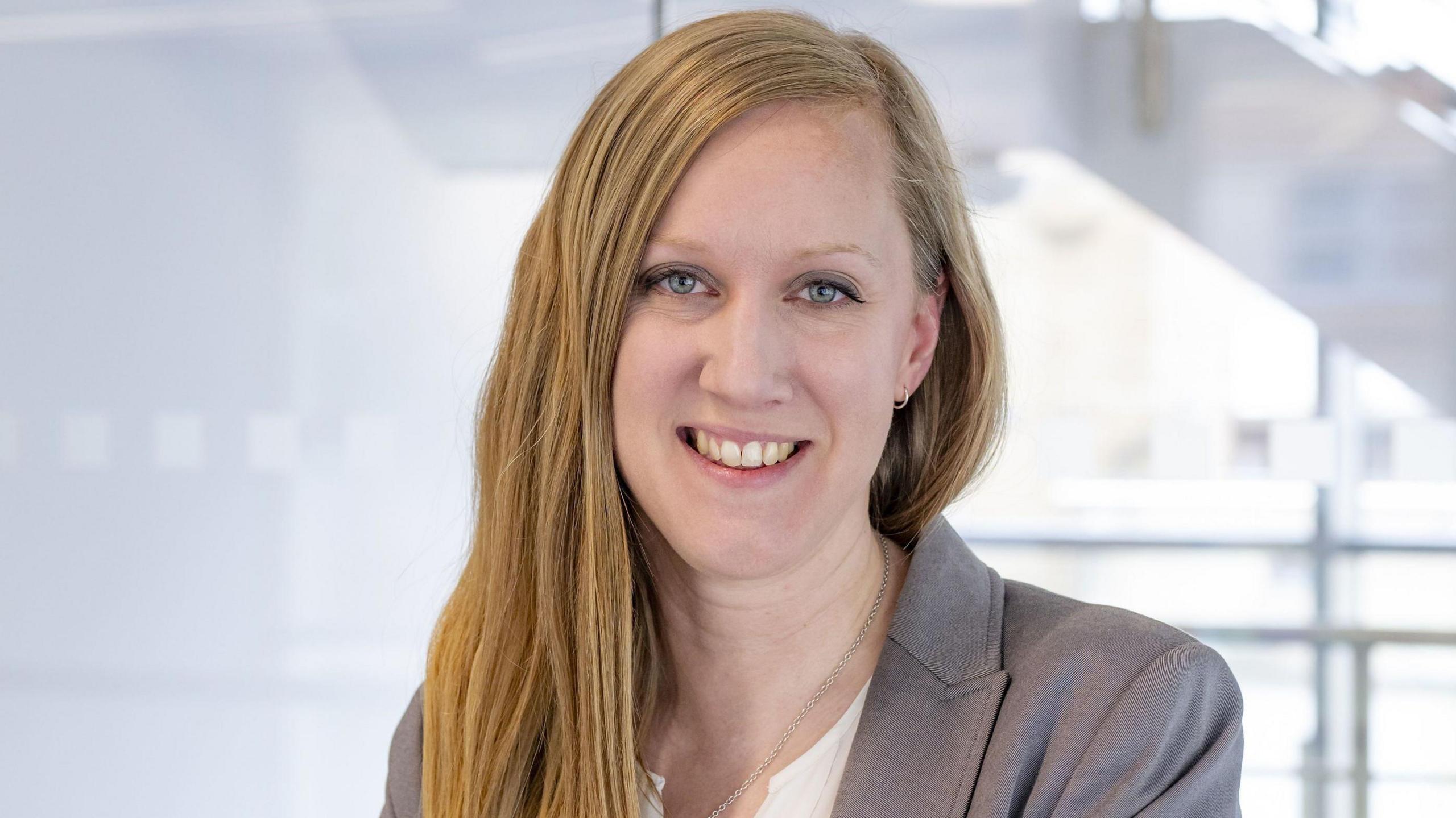 A publicity shot of a smiling Dr Victoria Goss. She has long blonde straight hair and is wearing a grey suit.