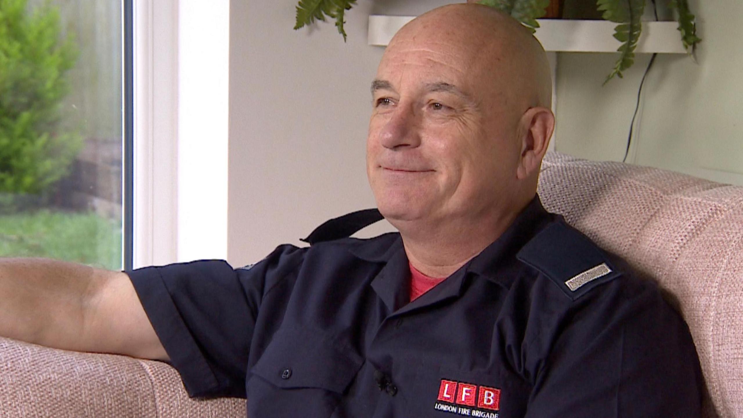 Andy Coote sits on his sofa looking into the middle distance, wearing his dark blue firefighter's uniform. 