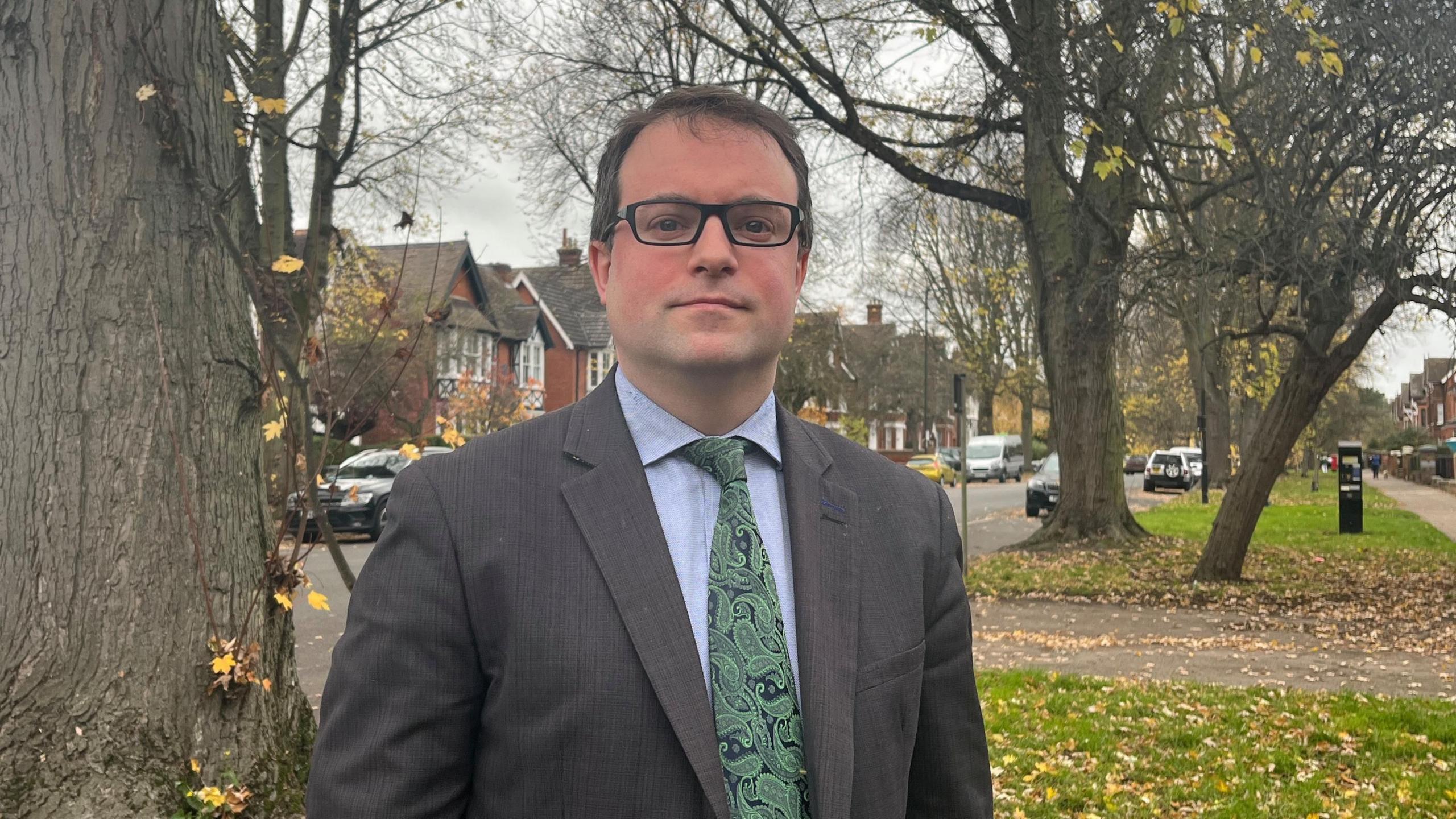 A man with glasses dressed in a suit standing by a road with lots of trees either side of it.