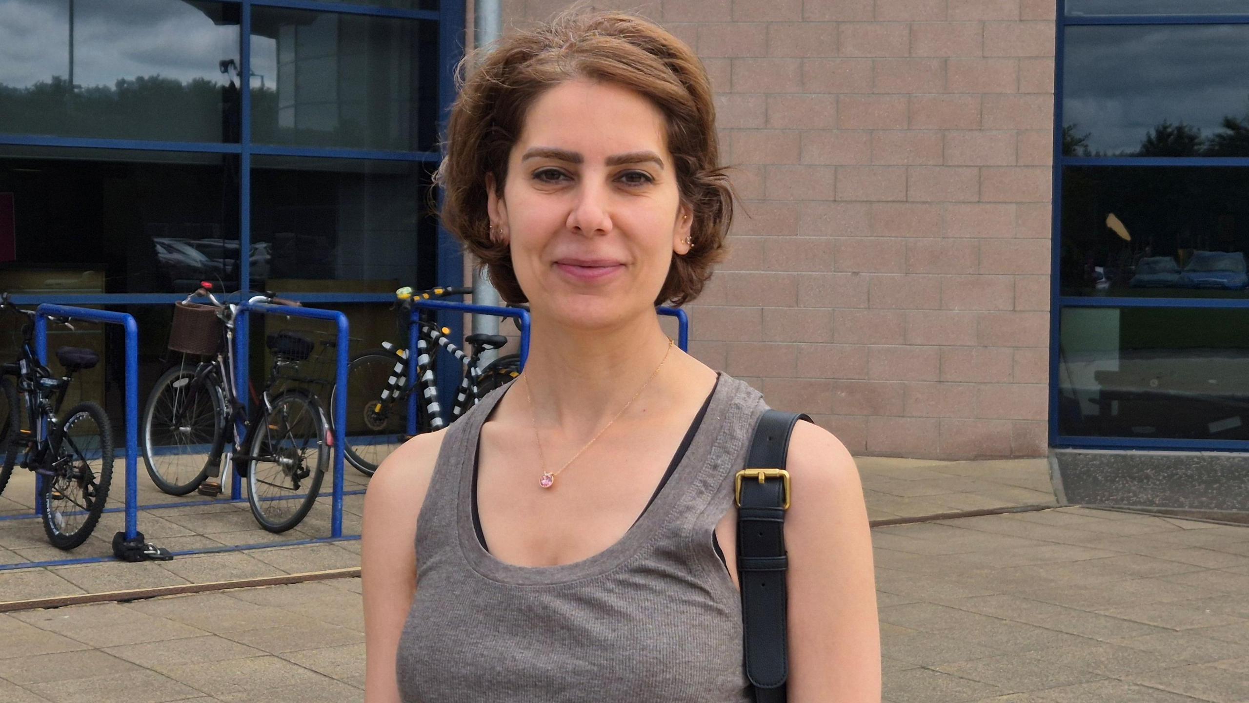 A woman with dark brown hair, wearing a grey top, standing outside a gym