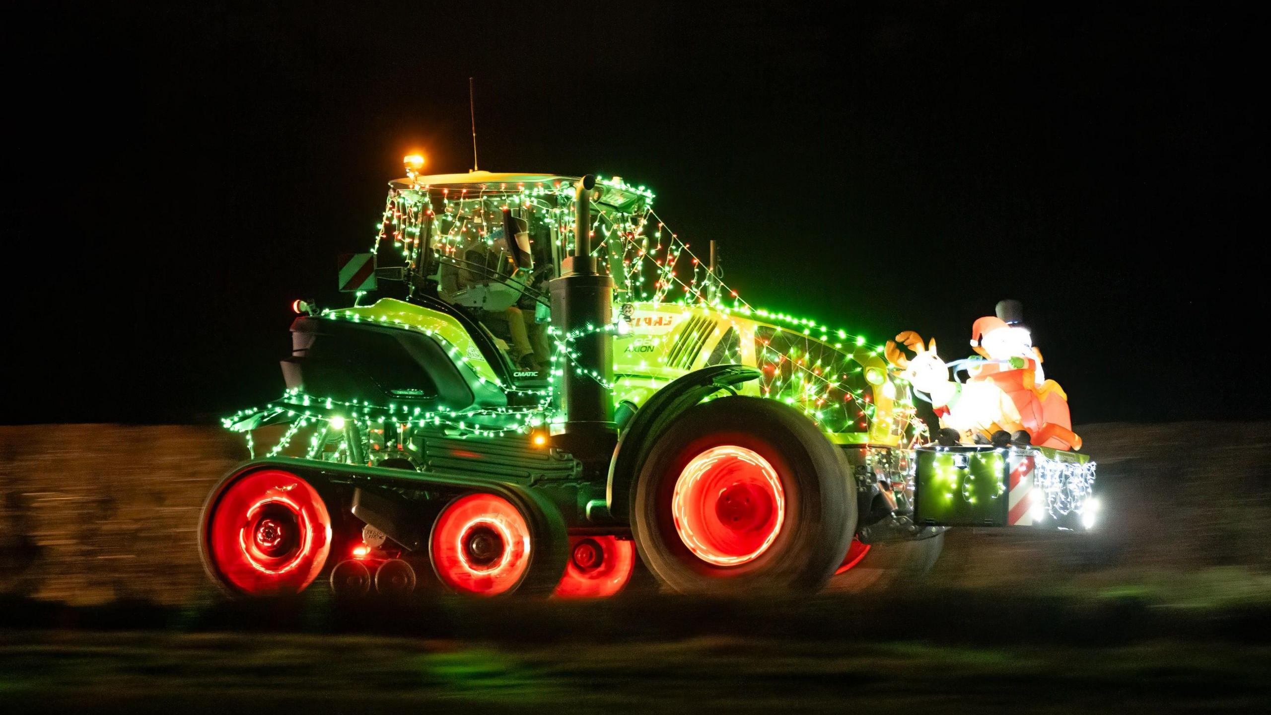 A tractor with its cab covered in fairy lights and its wheels shining red. In a box on its front there is a light up snowman, Santa and reindeer and the landscape is blurred as if the tractor is moving quickly.